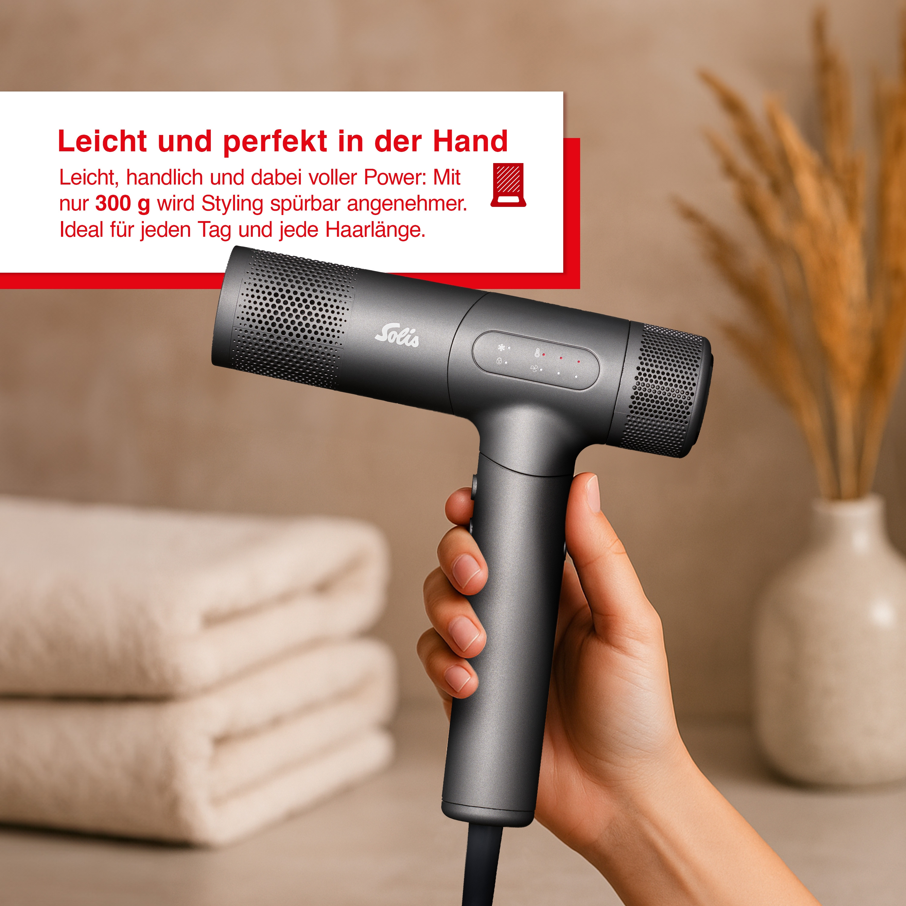 Eine Hand hält einen dunklen Haartrockner mit Solis-Logo. Daneben liegen Handtücher.