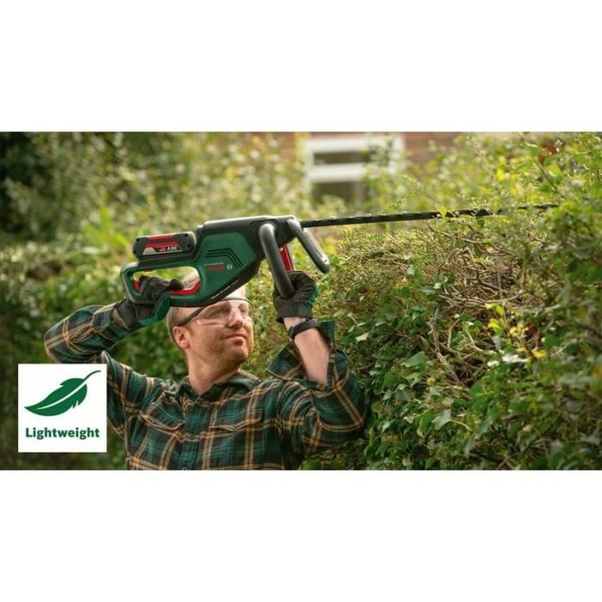 Mann trimmt Hecke mit grünem und schwarzem Hedge Trimmer, trägt kariertes Hemd.