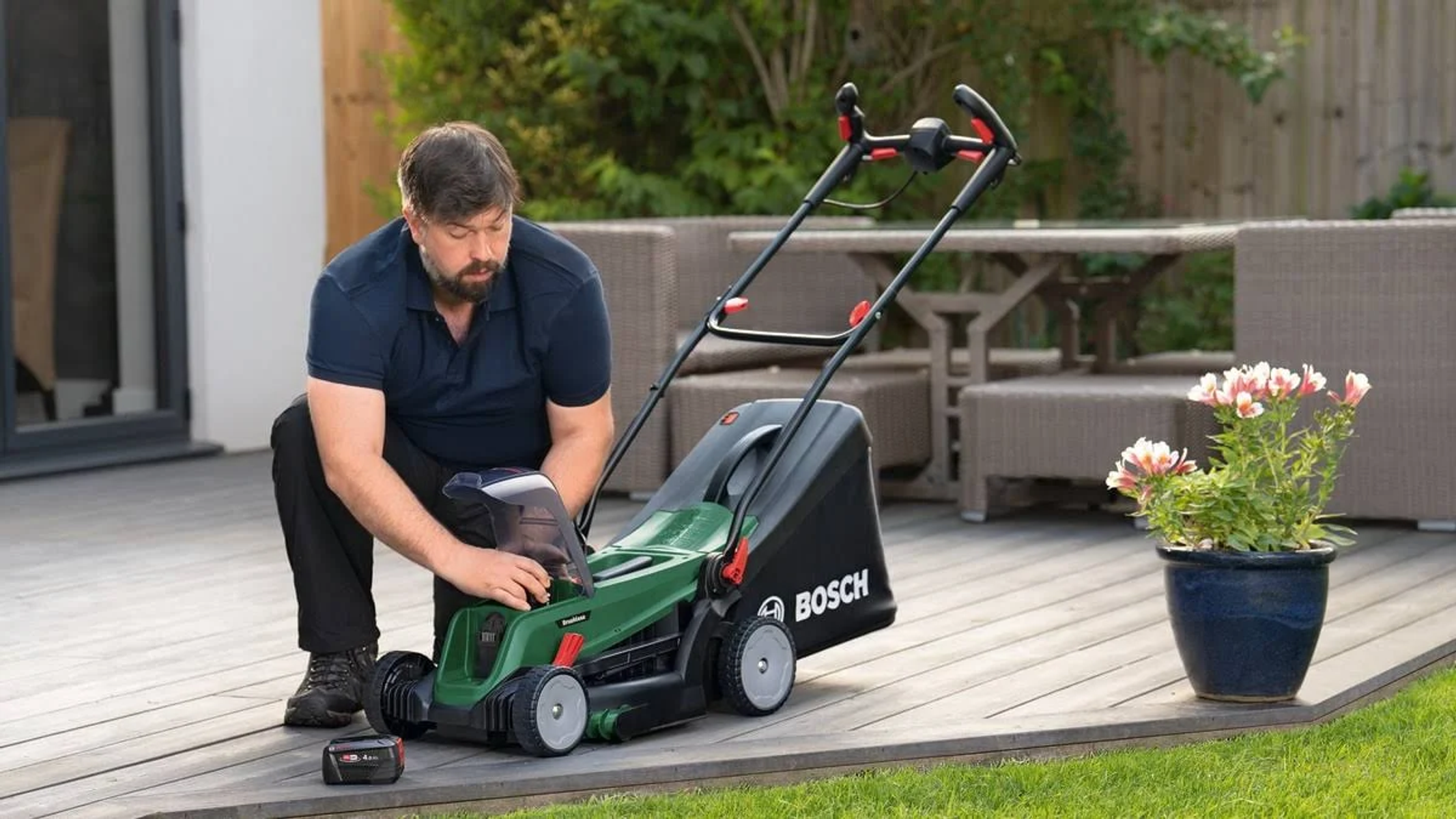 Mann kniet neben einem grünen Bosch Rasenmäher auf einer Holzterrasse.