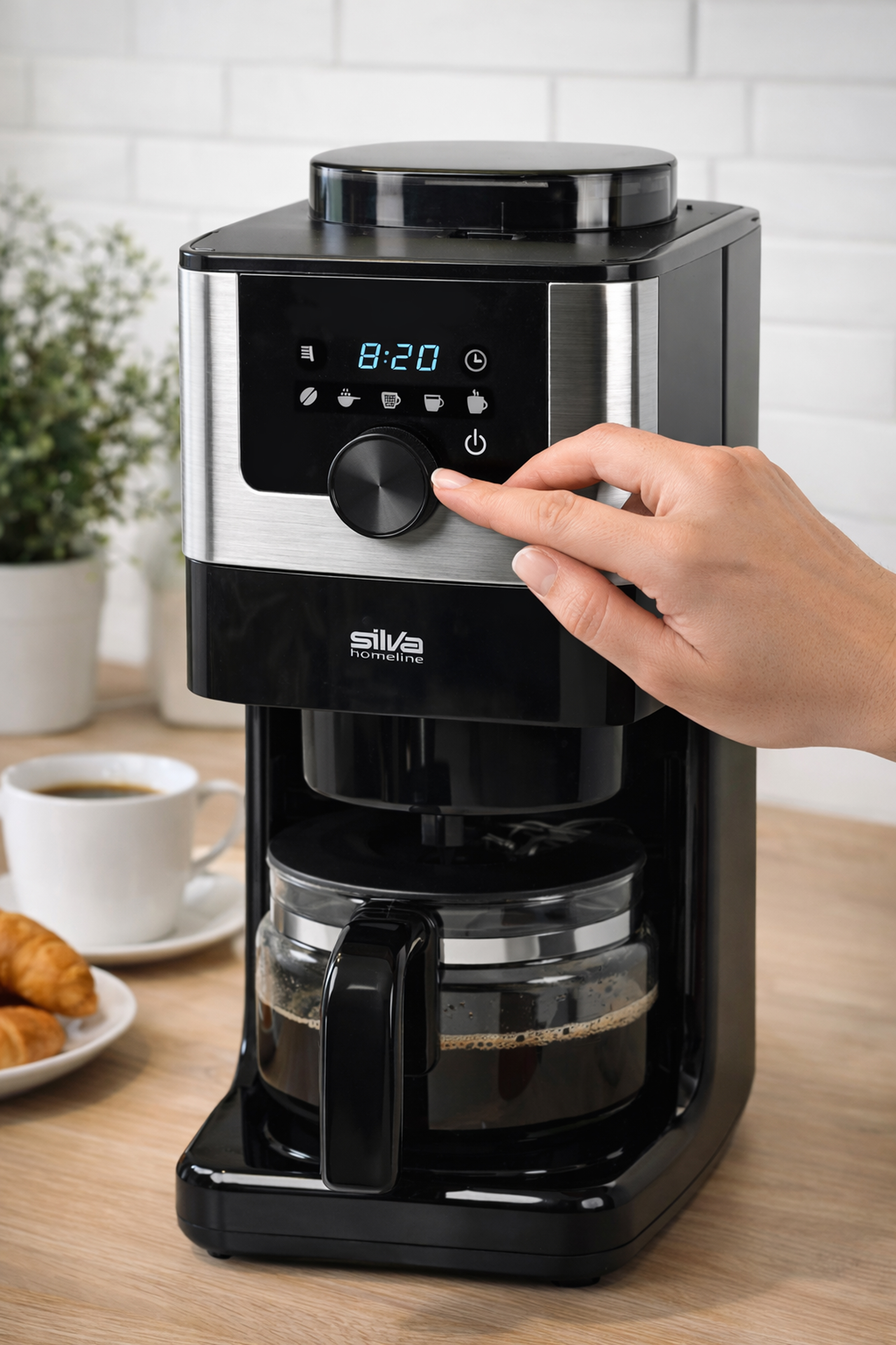 Eine Hand bedient eine Kaffeemaschine mit digitaler Anzeige. daneben stehen Kaffee und Croissants.