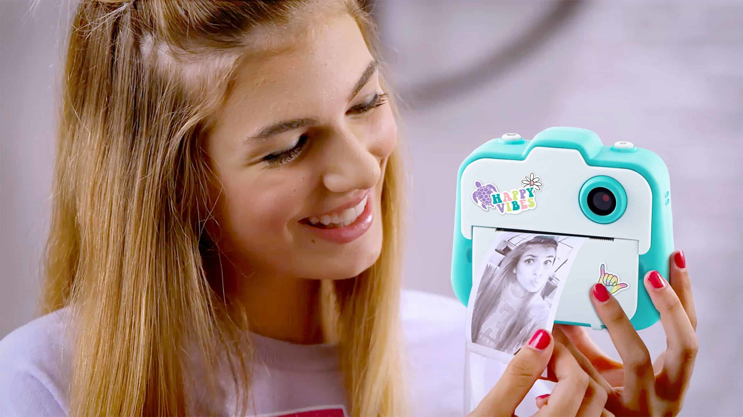 Una ragazza con i capelli biondi tiene una fotocamera istantanea turchese che stampa una foto in bianco e nero.