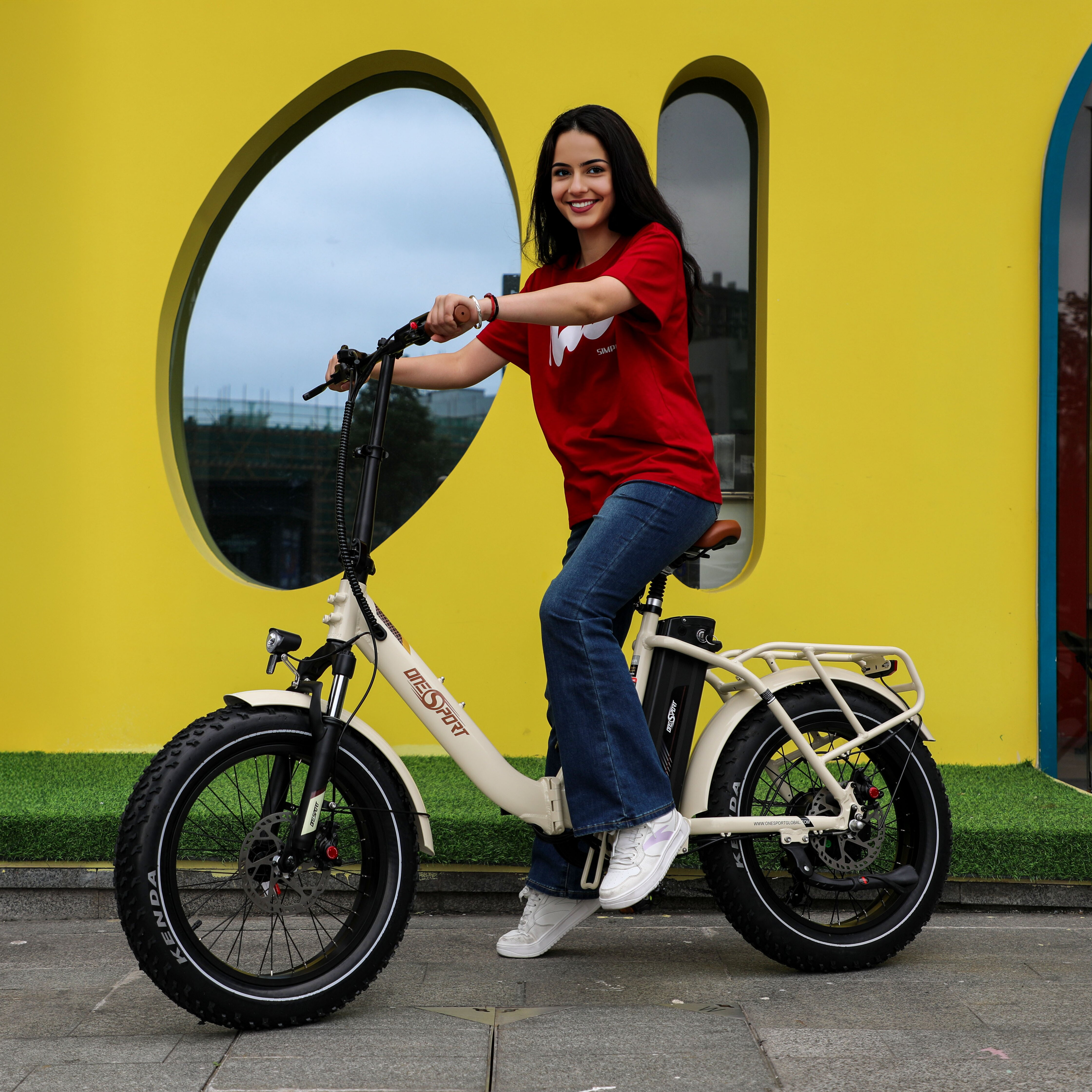 Una giovane donna in jeans e maglietta rossa siede su una e-bike color crema davanti a un muro giallo.