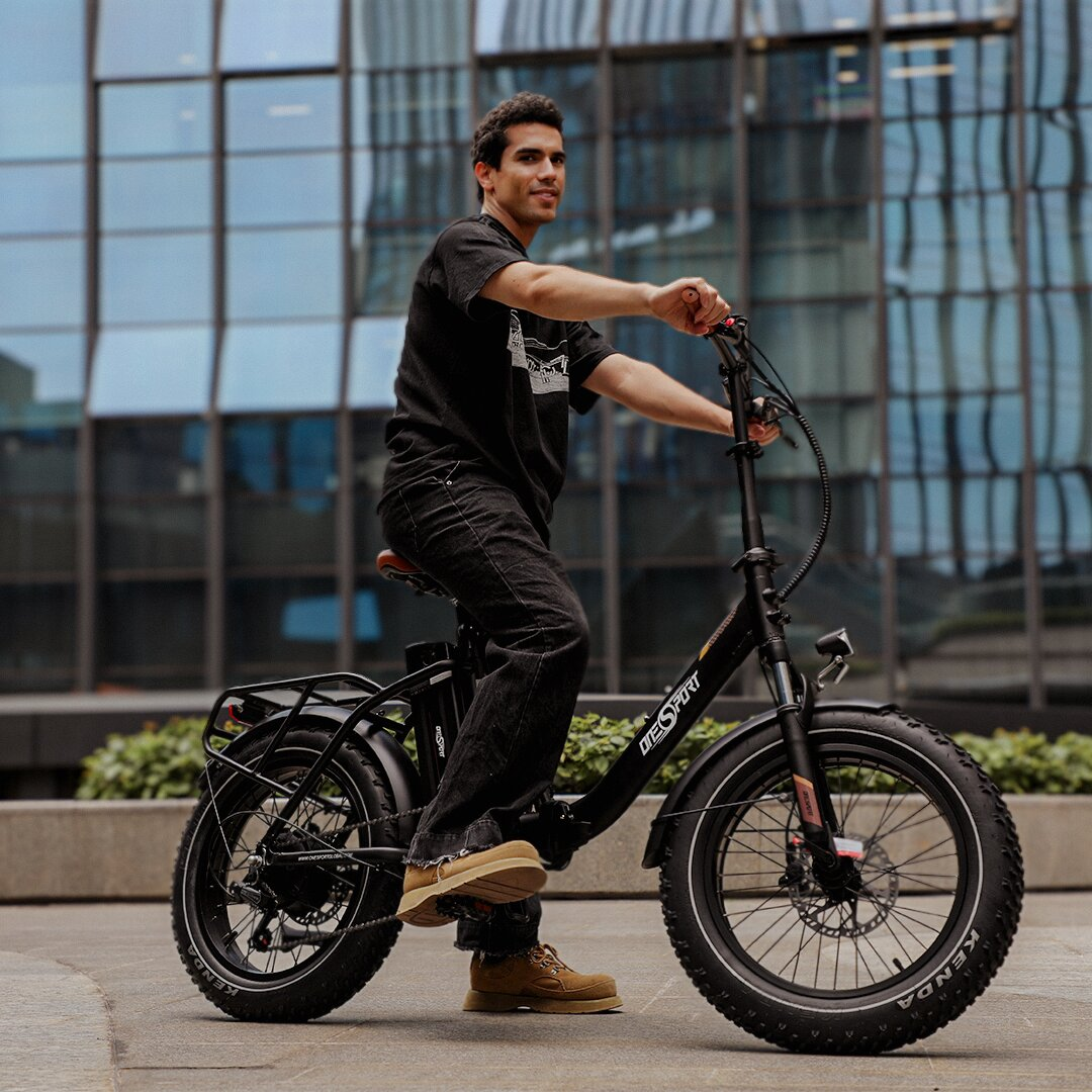 Uomo in maglietta nera e jeans guida e-bike con copertoni larghi davanti a edificio.