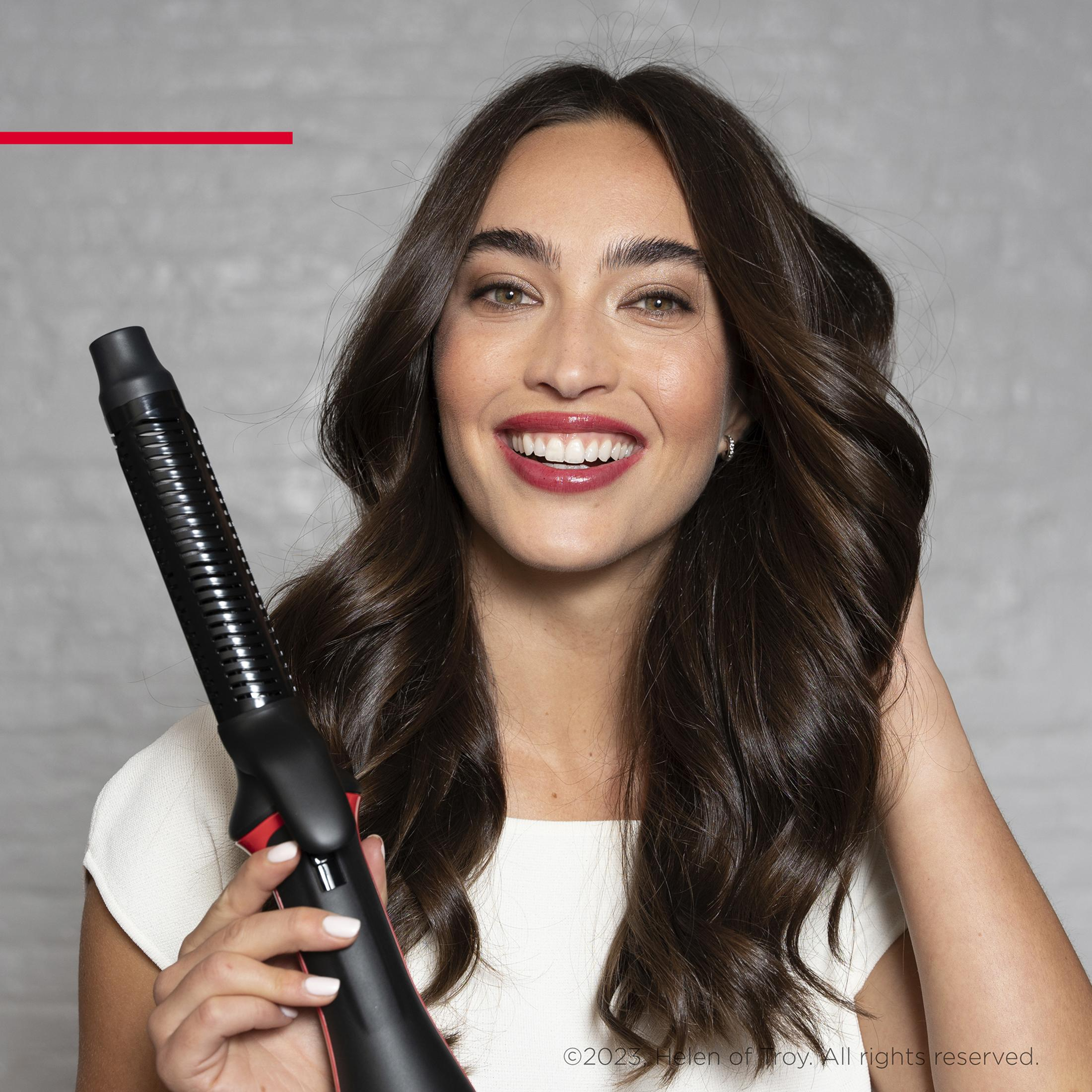 Een vrouw met lang bruin haar houdt een zwarte haardroger vast, lachend.