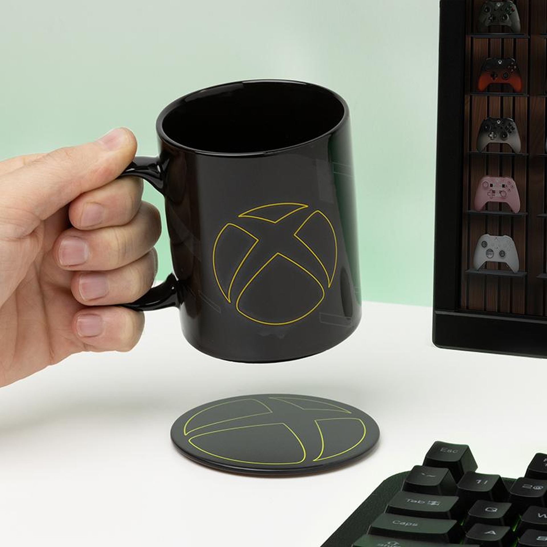 Czarny kubek Xbox z żółtym logo trzymany przez dłoń. W tle ekspozycja kontrolerów.