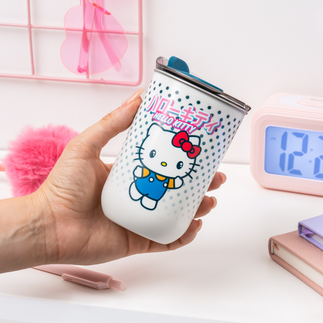 Dłoń trzyma biały kubek Hello Kitty z niebieskimi kropkami. W tle jest budzik.