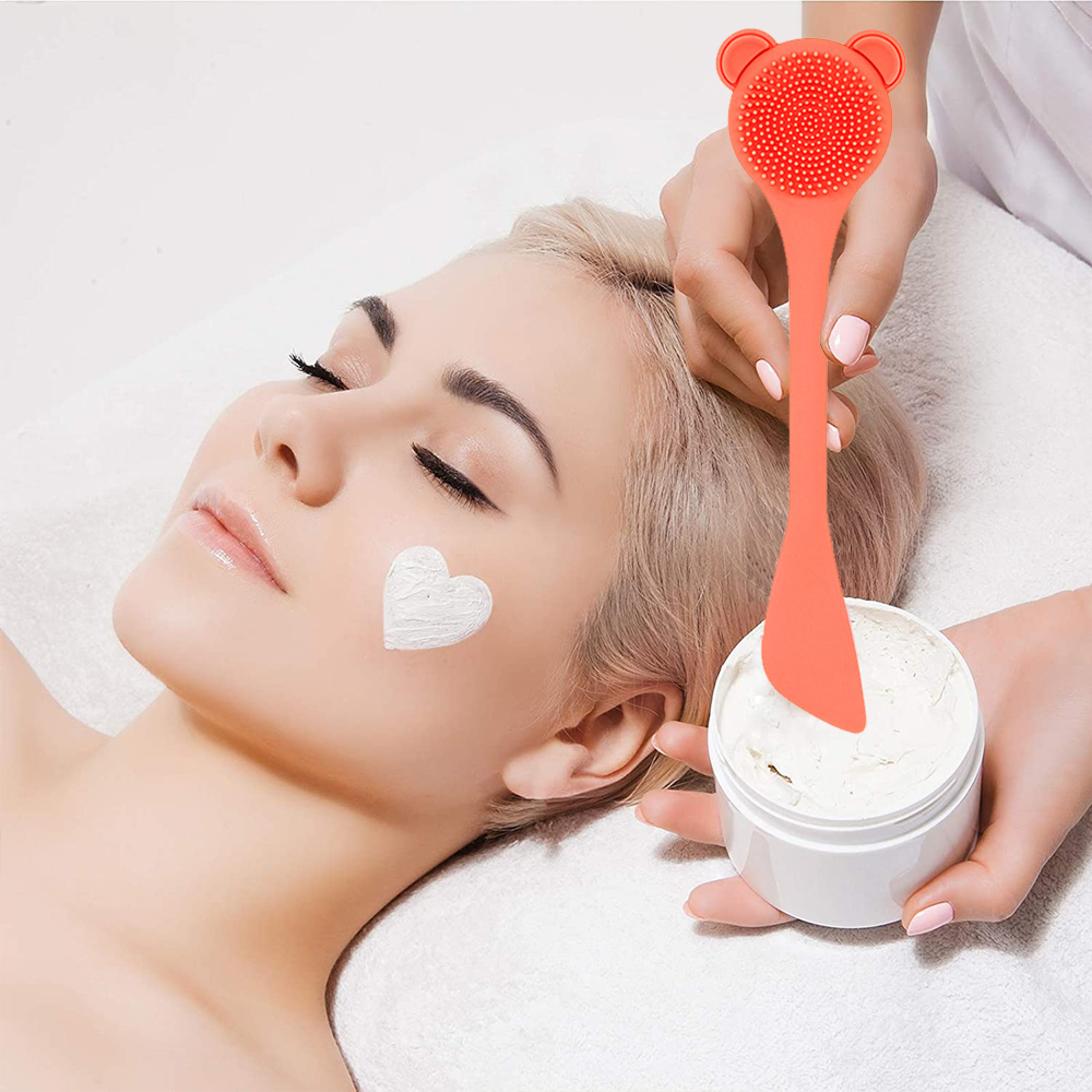 Eine Frau mit weißer Gesichtsmaske und einem orangefarbenen Werkzeug, das eine Creme aufträgt.