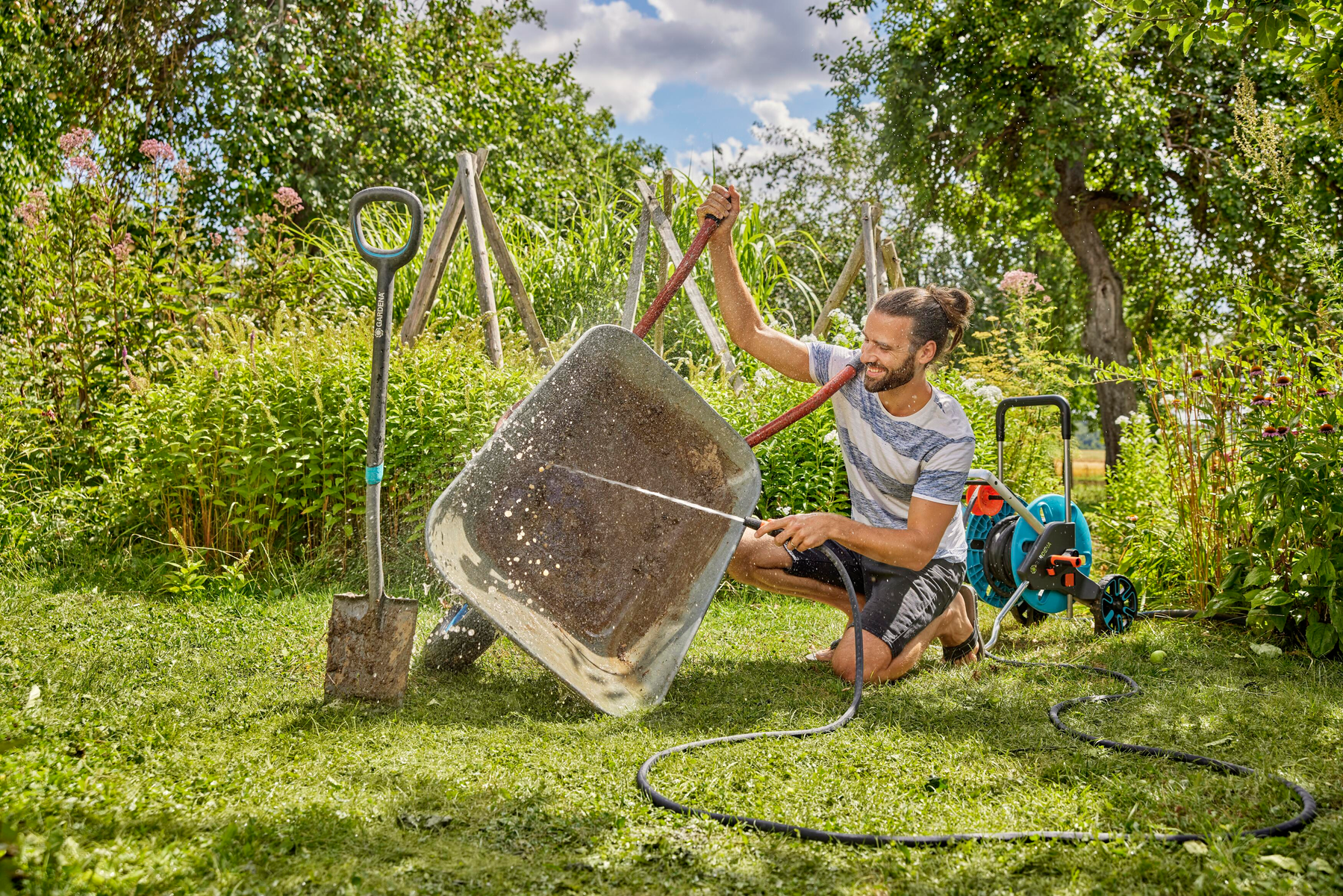 Uomo lava carriola con tubo da giardino, pala e avvolgitubo nelle vicinanze.
