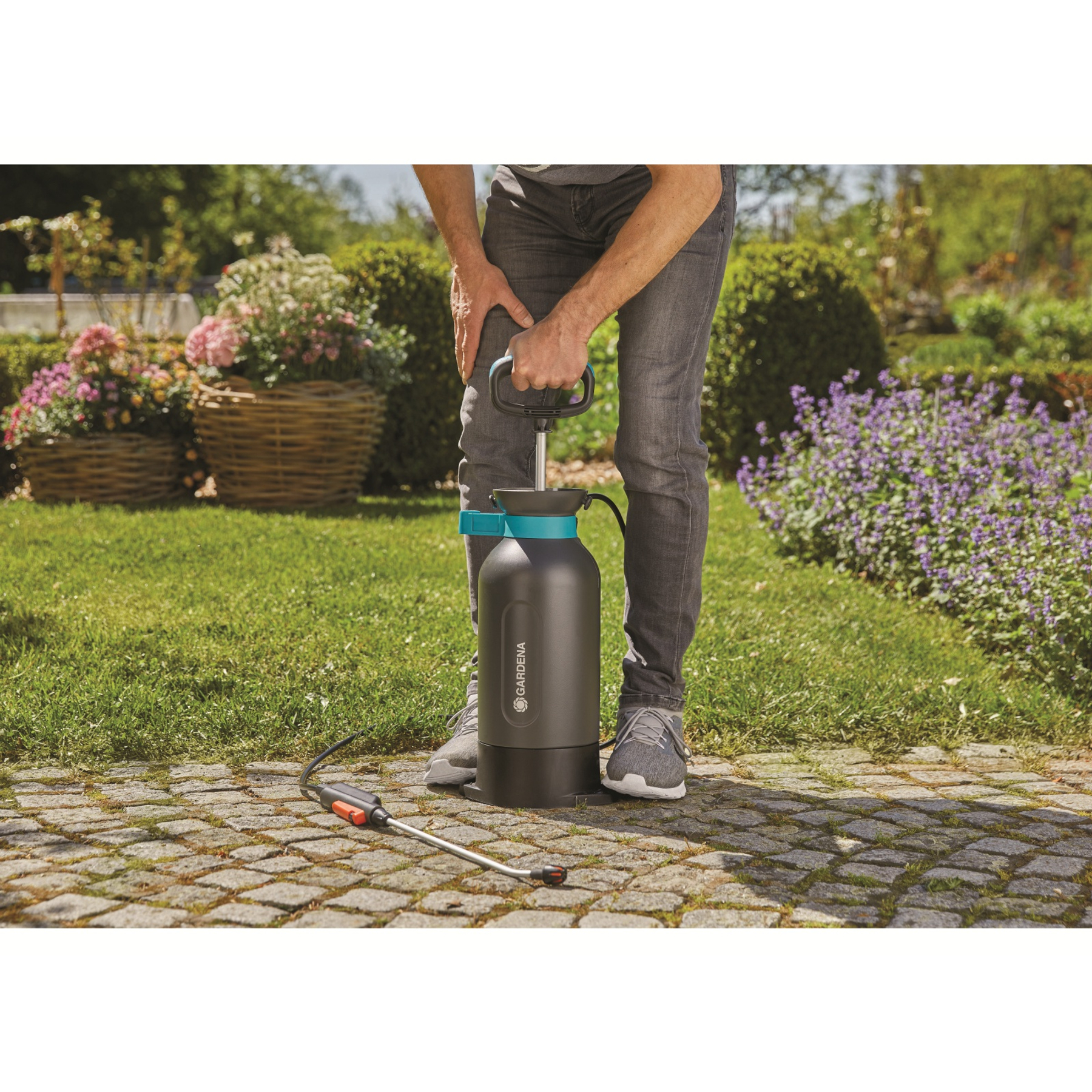 Uomo con spruzzatore su un sentiero lastricato in un giardino con fiori e cespugli.