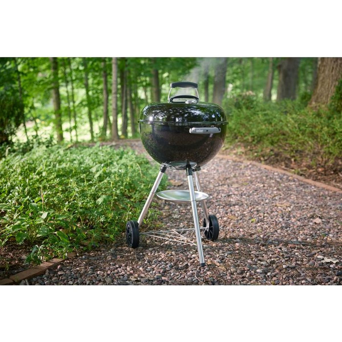 Ein schwarzer Weber Kugelgrill steht auf einem Schotterweg, umgeben von grüner Vegetation und Bäumen.