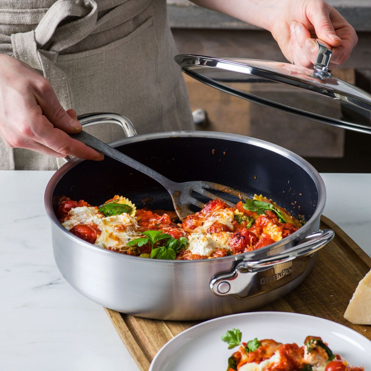 Eine Person hebt den Deckel von einem Topf mit Pasta, Tomaten und Käse.