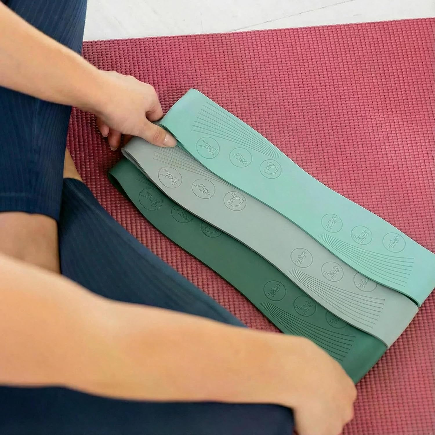 Drei Widerstandsbänder in Pastellfarben liegen auf einer pinken Yogamatte. Beine einer Person sind sichtbar.