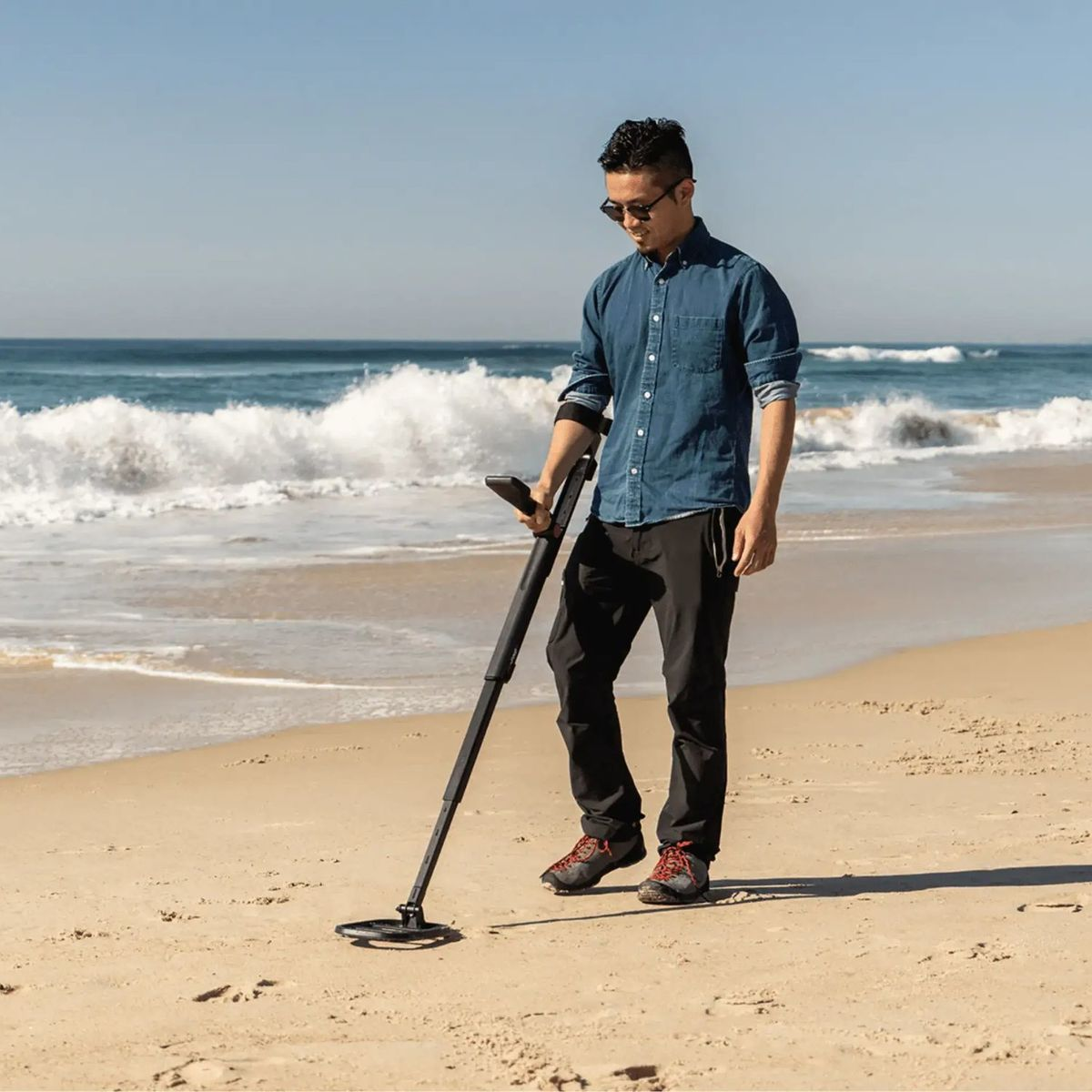 Mann mit Metalldetektor am Strand, Wellen im Hintergrund.