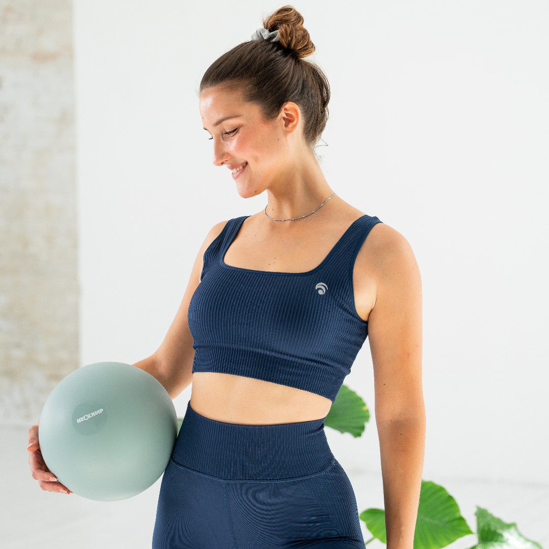 Eine Frau im marineblauen gerippten Sport-BH und Leggings hält einen hellblauen Gymnastikball.