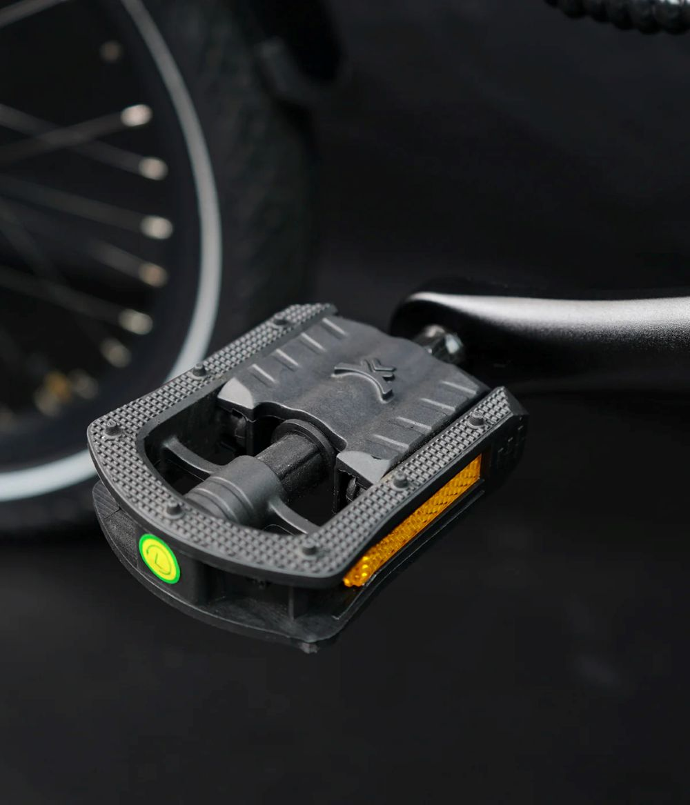 Pedal de bicicleta negro con reflectores y un acento verde, junto a una rueda.