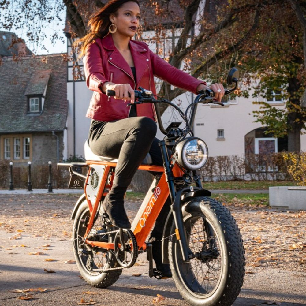 Una mujer monta una bicicleta eléctrica naranja en una calle con hojas caídas.