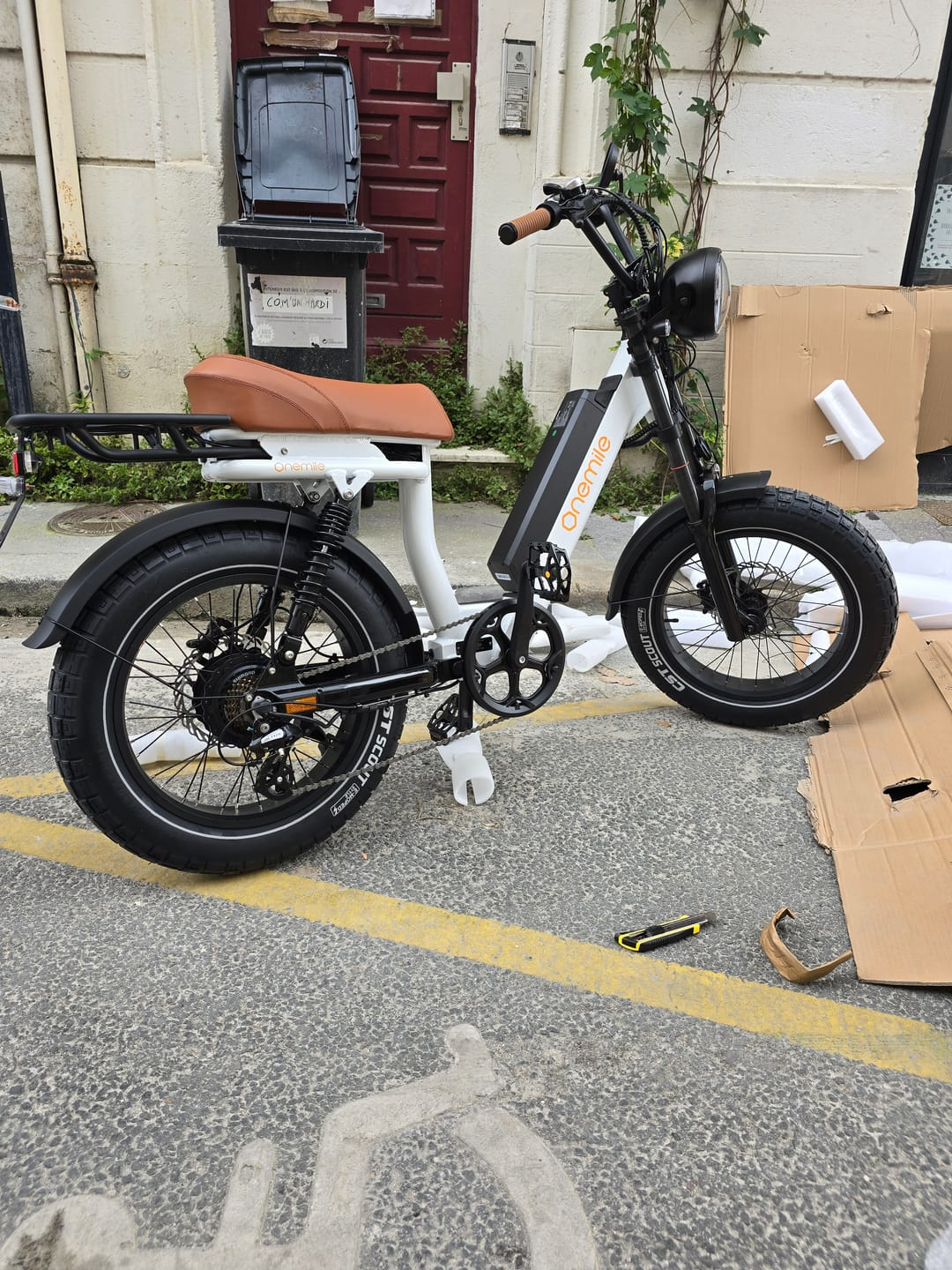 Una e-bike bianca con sella marrone è parcheggiata sul marciapiede di fronte a un edificio.