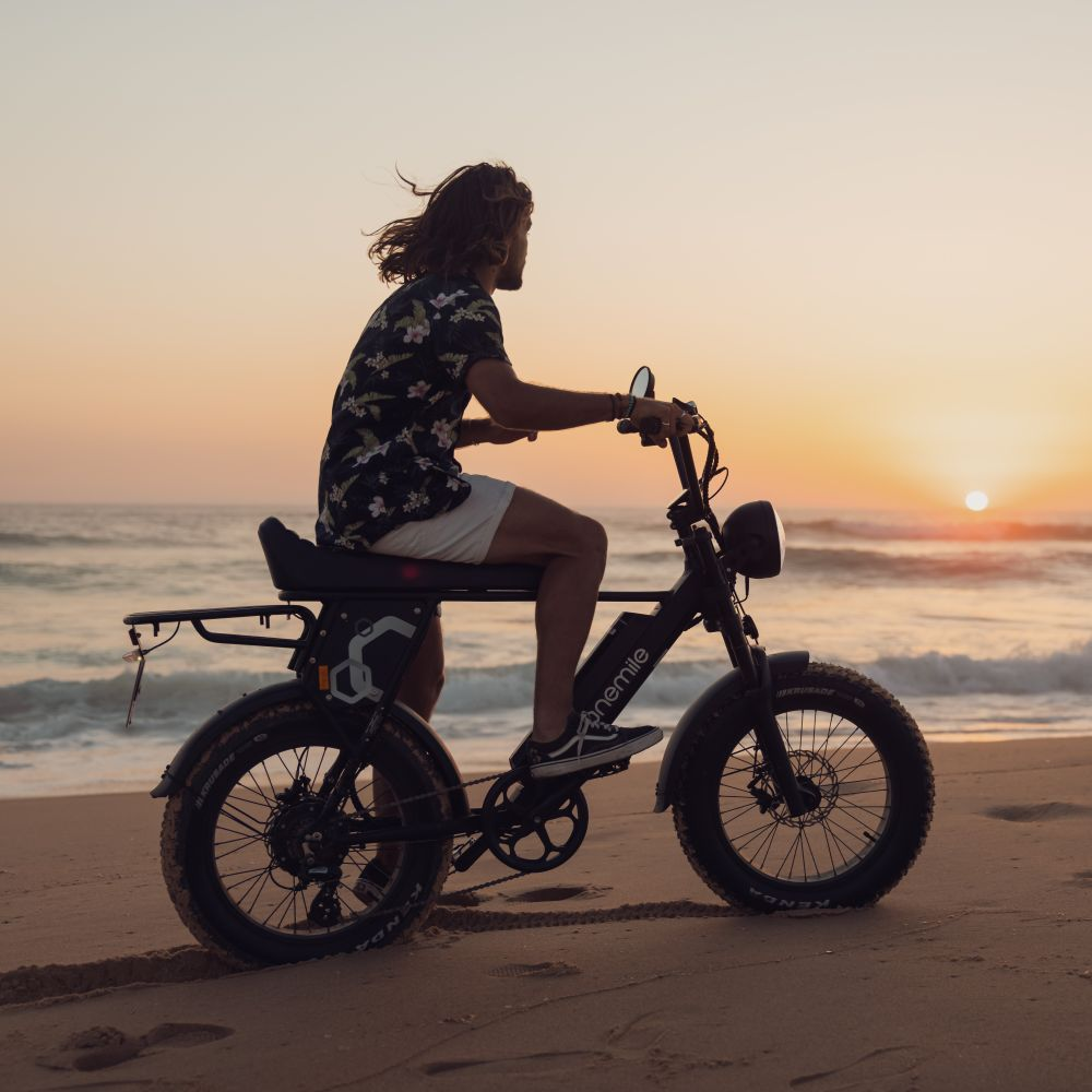 Hombre con pelo largo en bici eléctrica en playa al atardecer. Olas y cielo.
