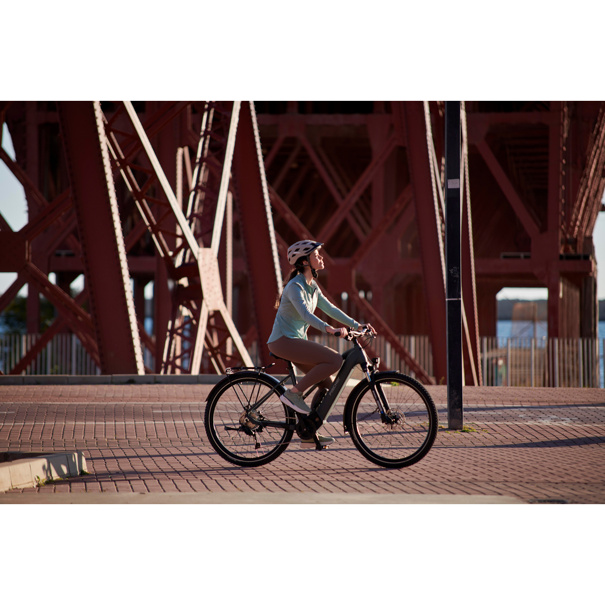 Eine Frau fährt mit einem E-Bike unter einer roten Stahlbrücke auf einem Kopfsteinpflasterweg.