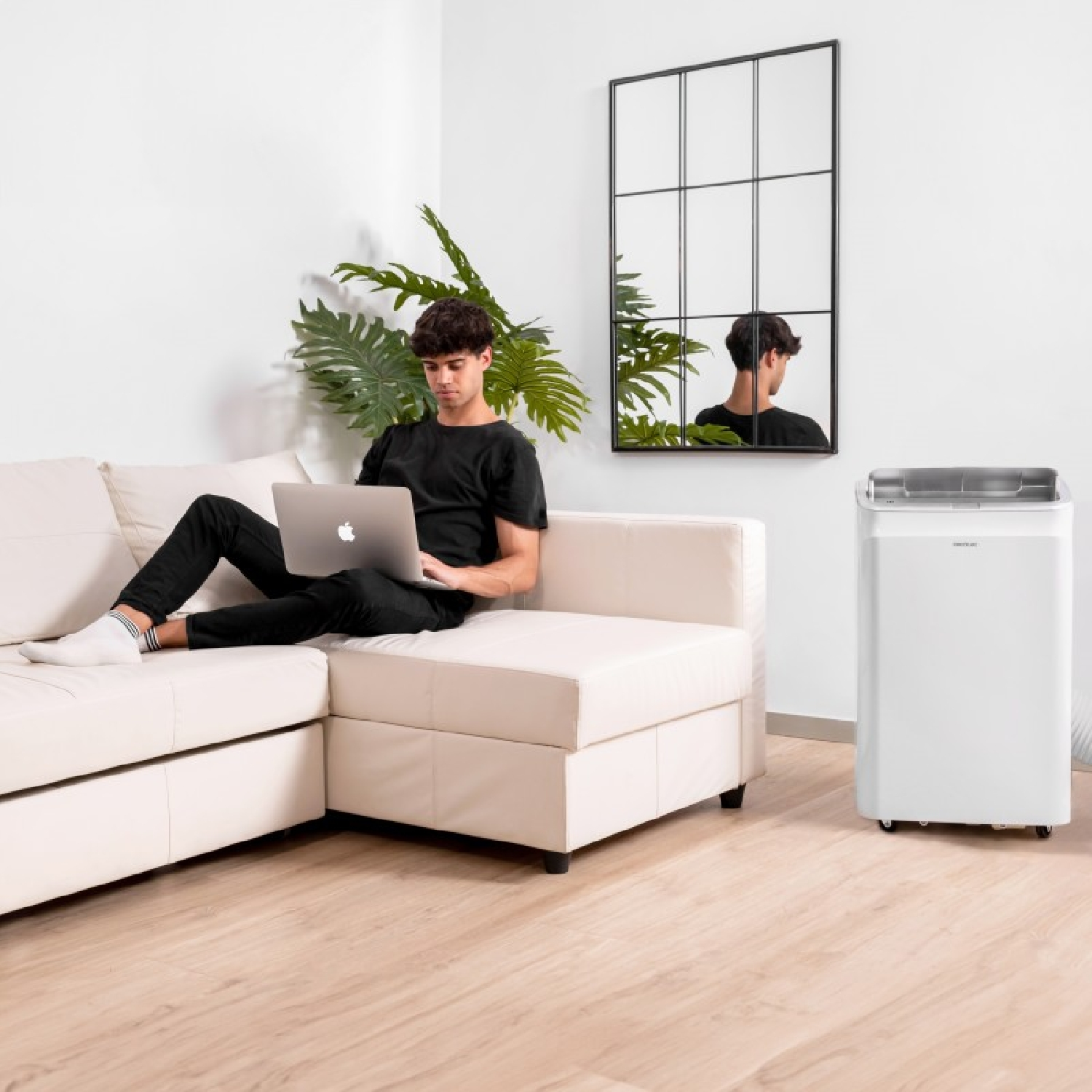Un uomo siede su un divano bianco usando un laptop. Un condizionatore portatile è nelle vicinanze.