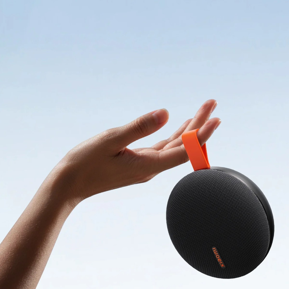Una mano tiene un altoparlante nero e rotondo con cinturino arancione contro il cielo.