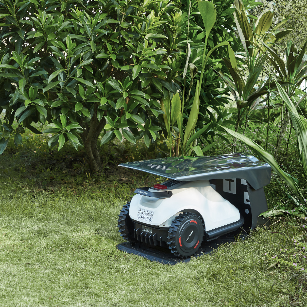 A white robotic lawn mower sits on grass under a tree and lush plants.