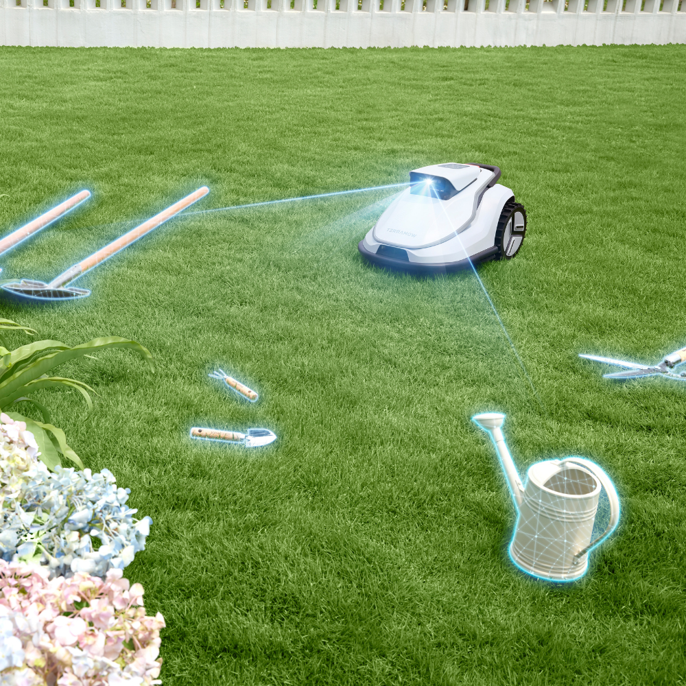 A white robotic lawn mower on grass with garden tools and a white garden fence.