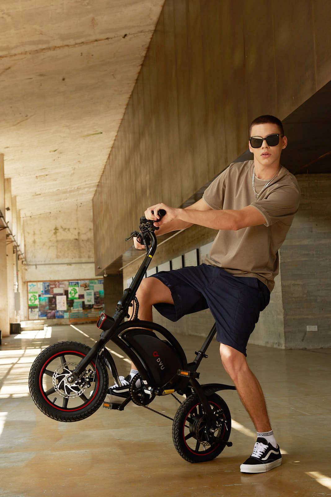 Hombre con gafas de sol en bici eléctrica negra, una rueda arriba, edificio de hormigón.