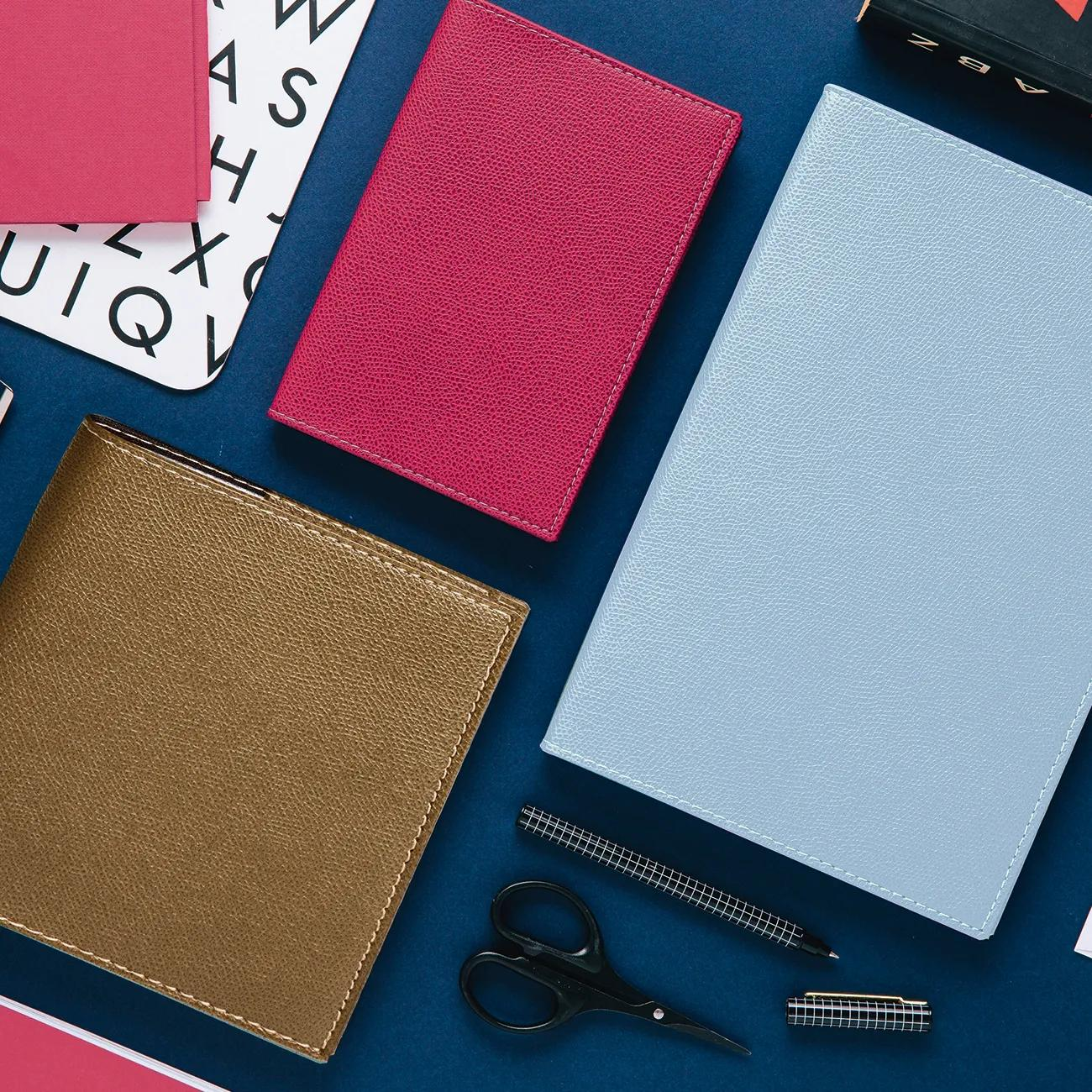 Tres cuadernos en rosa, azul y dorado con tijeras y bolígrafo sobre fondo azul.