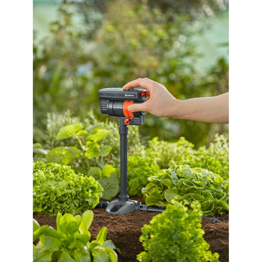 Una mano sujeta un aspersor de jardín negro y naranja entre plantas de lechuga.
