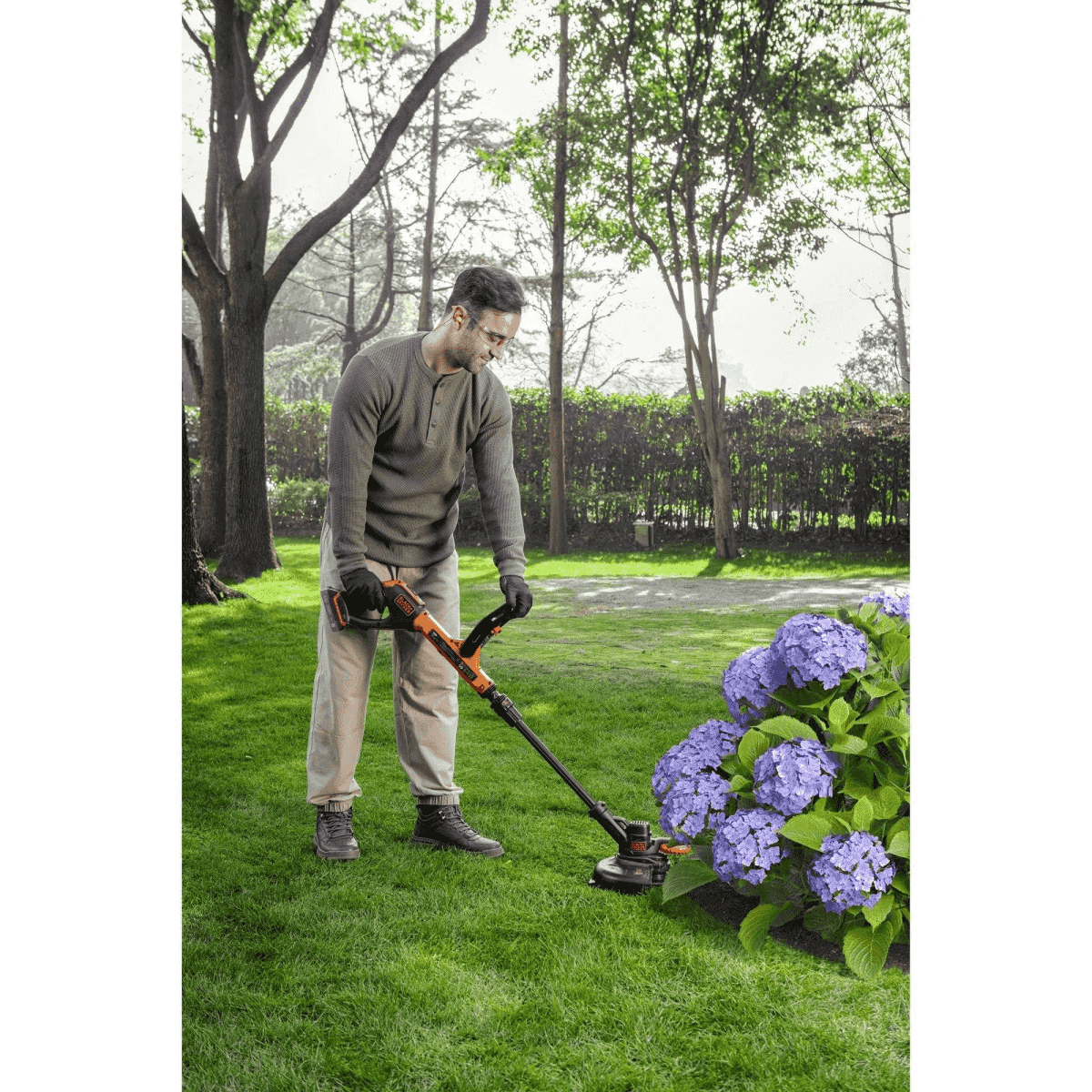 Un hombre recorta el césped con un cortabordes junto a hortensias azules en un jardín.