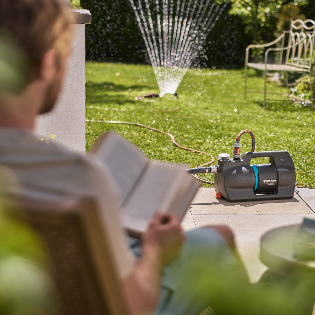 Uomo legge un libro in giardino mentre una pompa annaffia il prato.