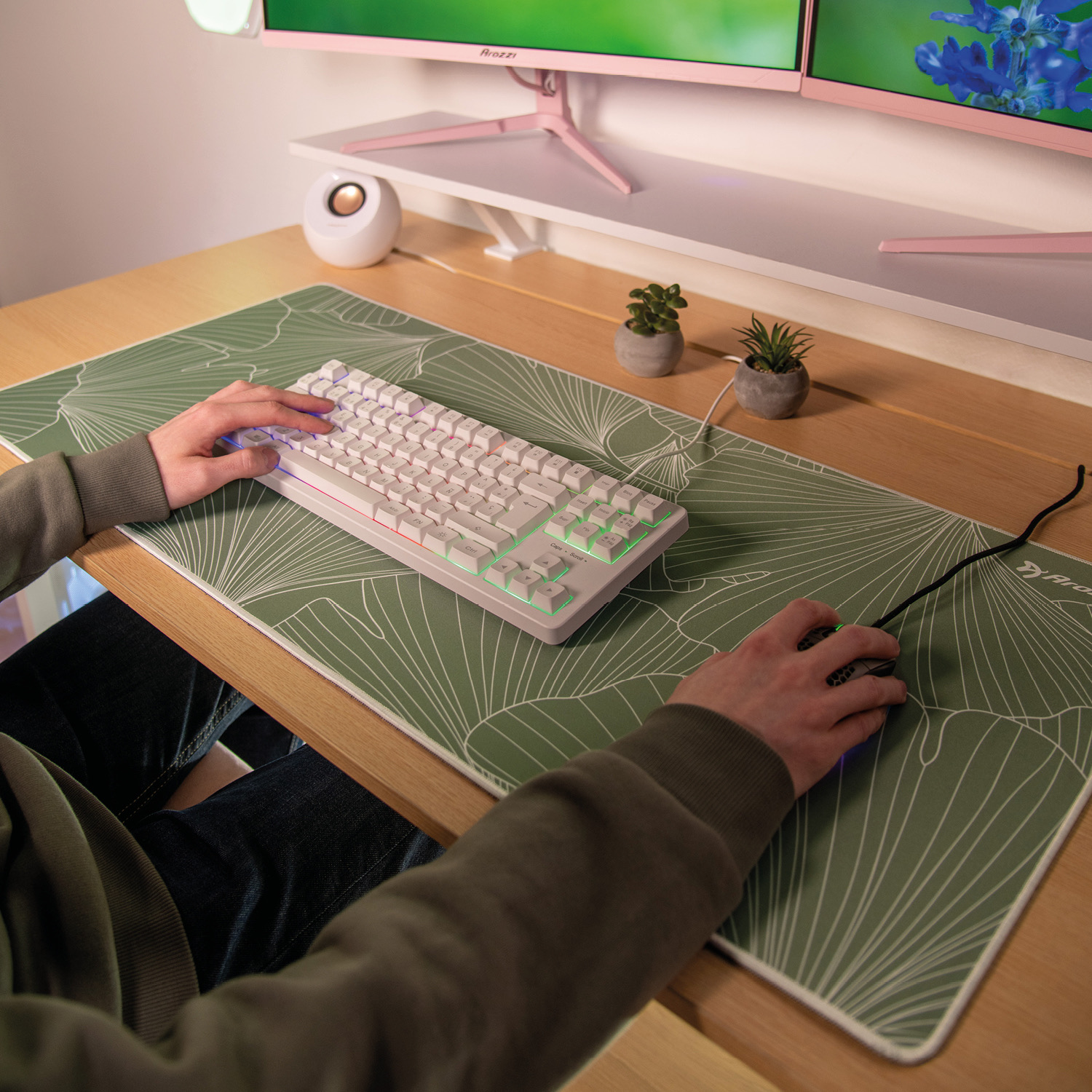 Hände auf einer weißen Tastatur und Maus auf einer grünen Matte, mit Monitoren im Hintergrund.