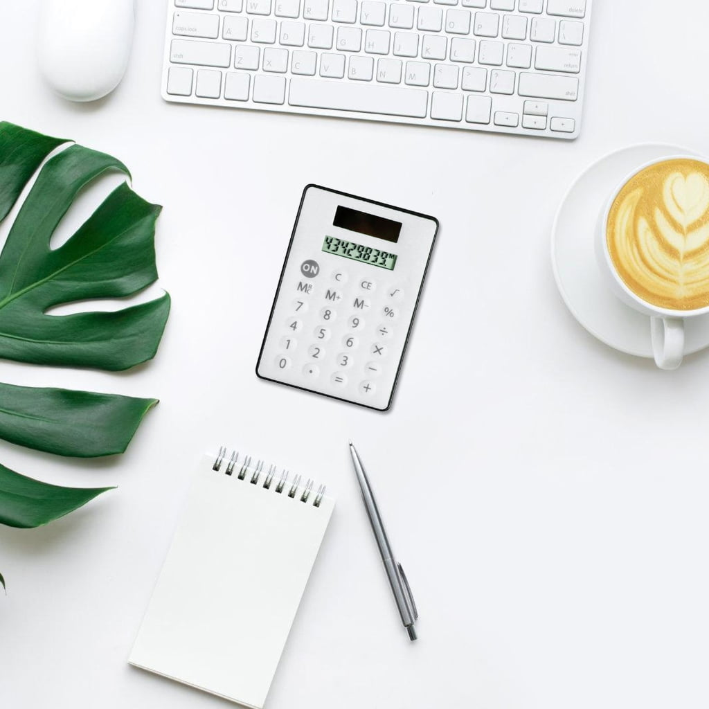 Eine flache Aufnahme eines Schreibtisches mit Taschenrechner, Kaffee, Notizbuch, Stift und Tastatur.