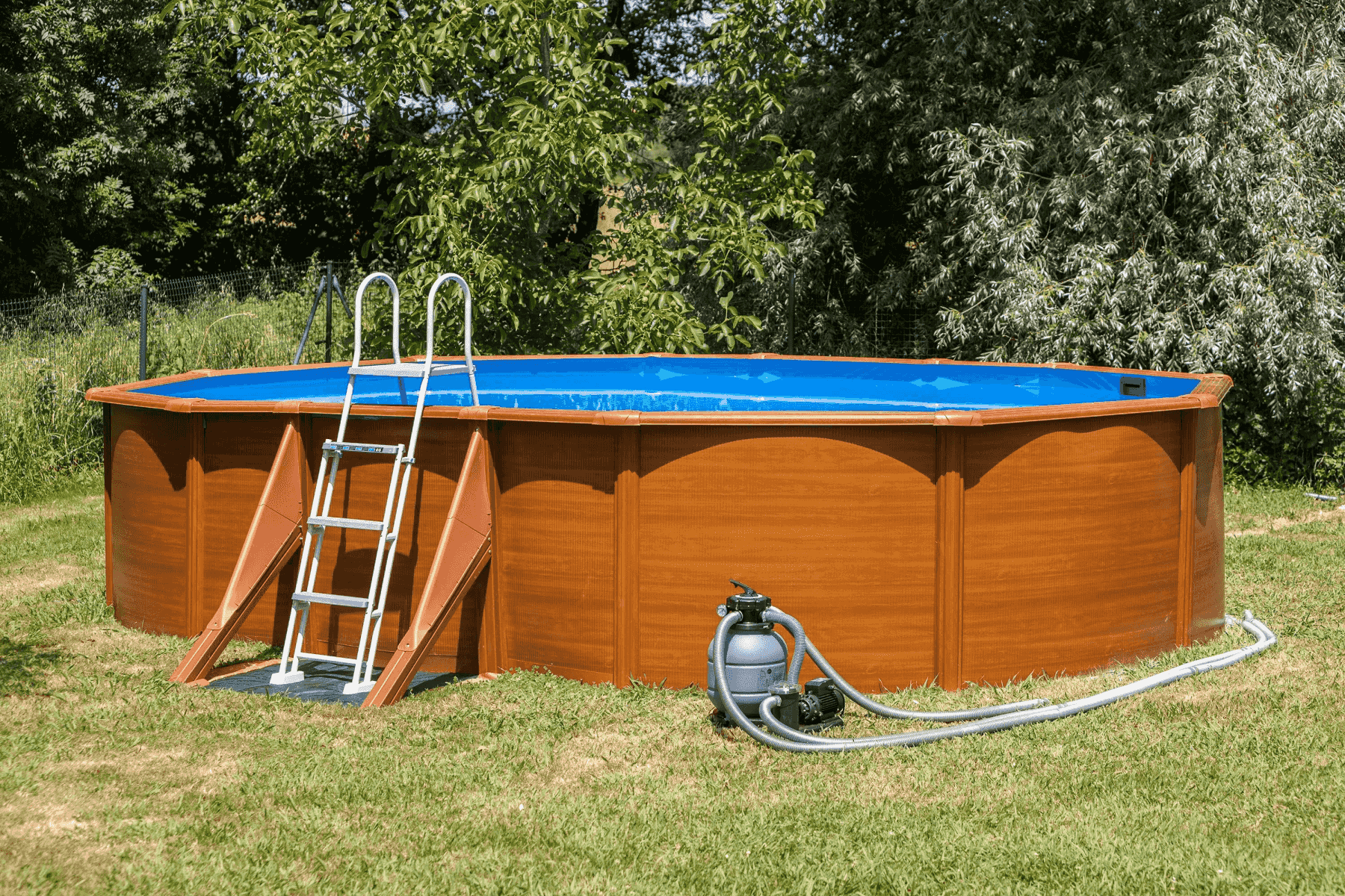 Una piscina ovalada con paredes de madera marrón y liner azul, con escalera y bomba en el jardín.