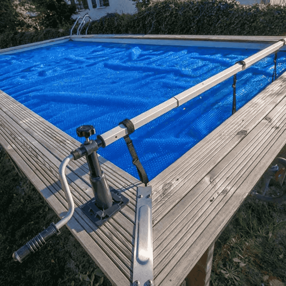 Una piscina con cubierta azul y sistema de enrollado plateado en una terraza de madera.