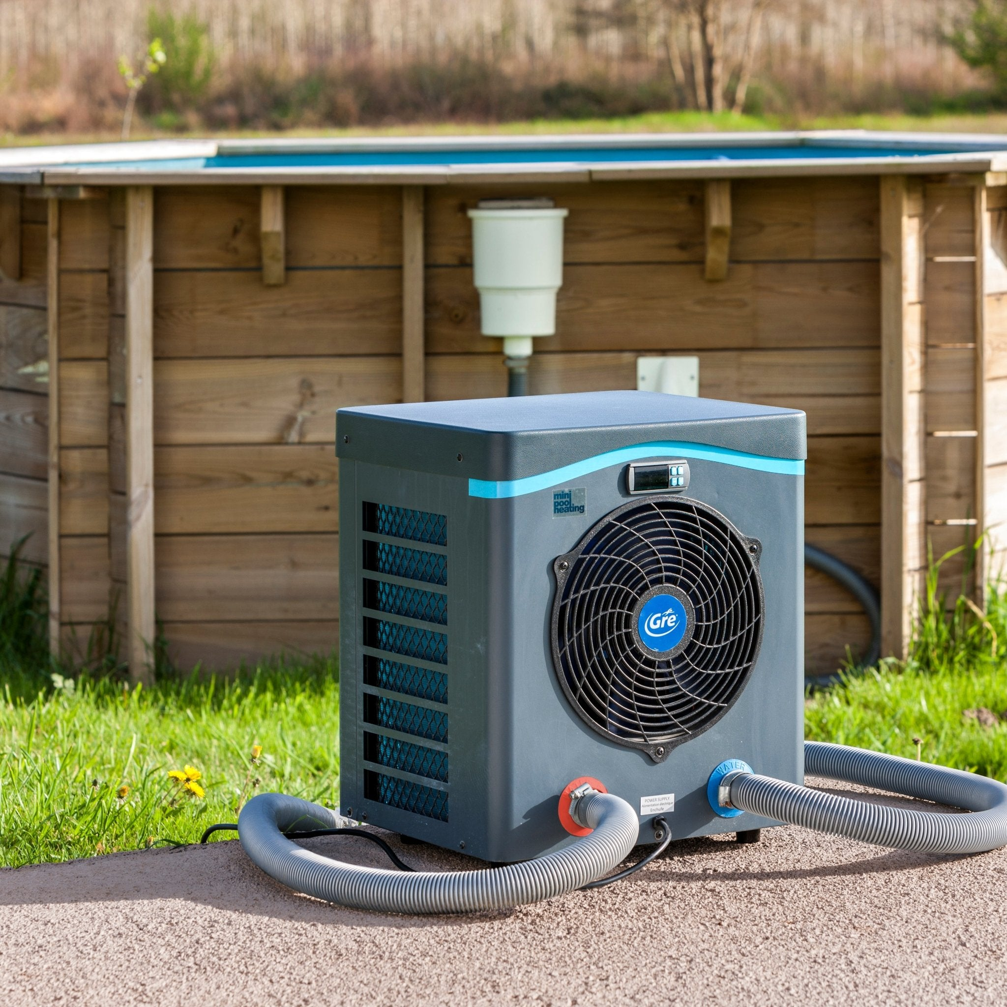 Una unidad gris de bomba de calor para piscina con ventilador se asienta sobre grava frente a una piscina de madera.