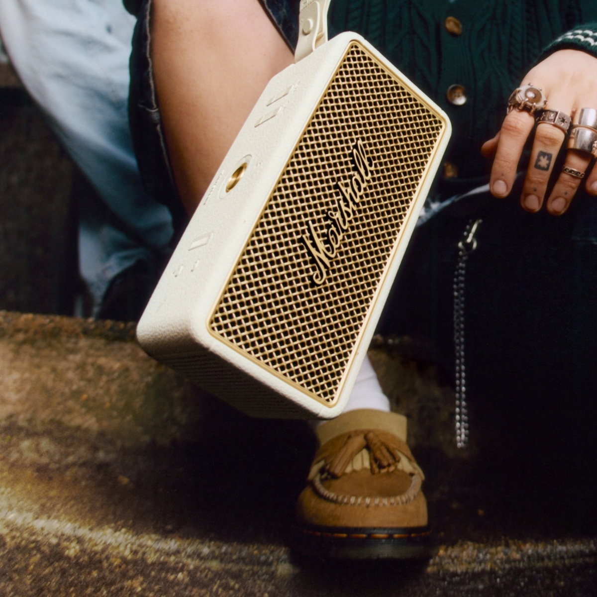 Eine Person hält einen weißen Marshall-Lautsprecher mit goldenem Grill. Sie trägt Ringe und Mokassins.