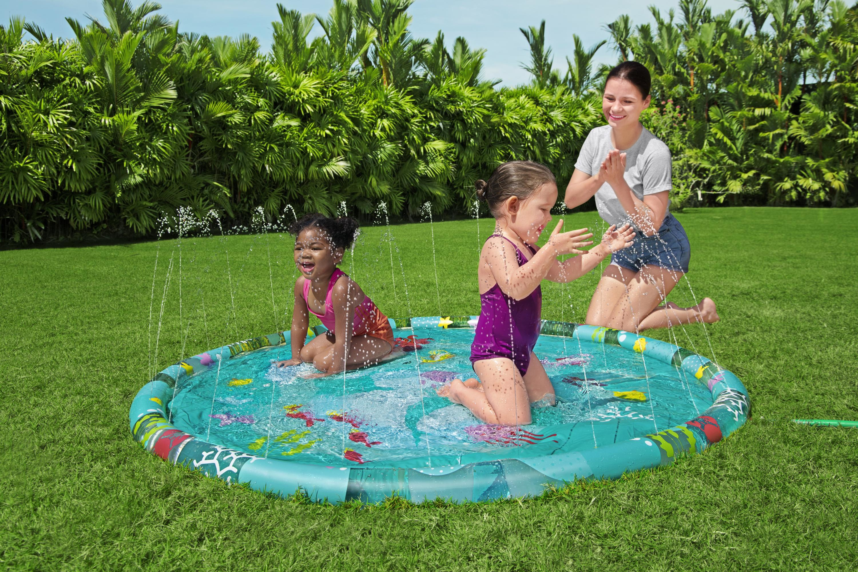 Dos niñas juegan en una piscina con aspersores mientras una mujer observa. Plantas verdes exuberantes las rodean.