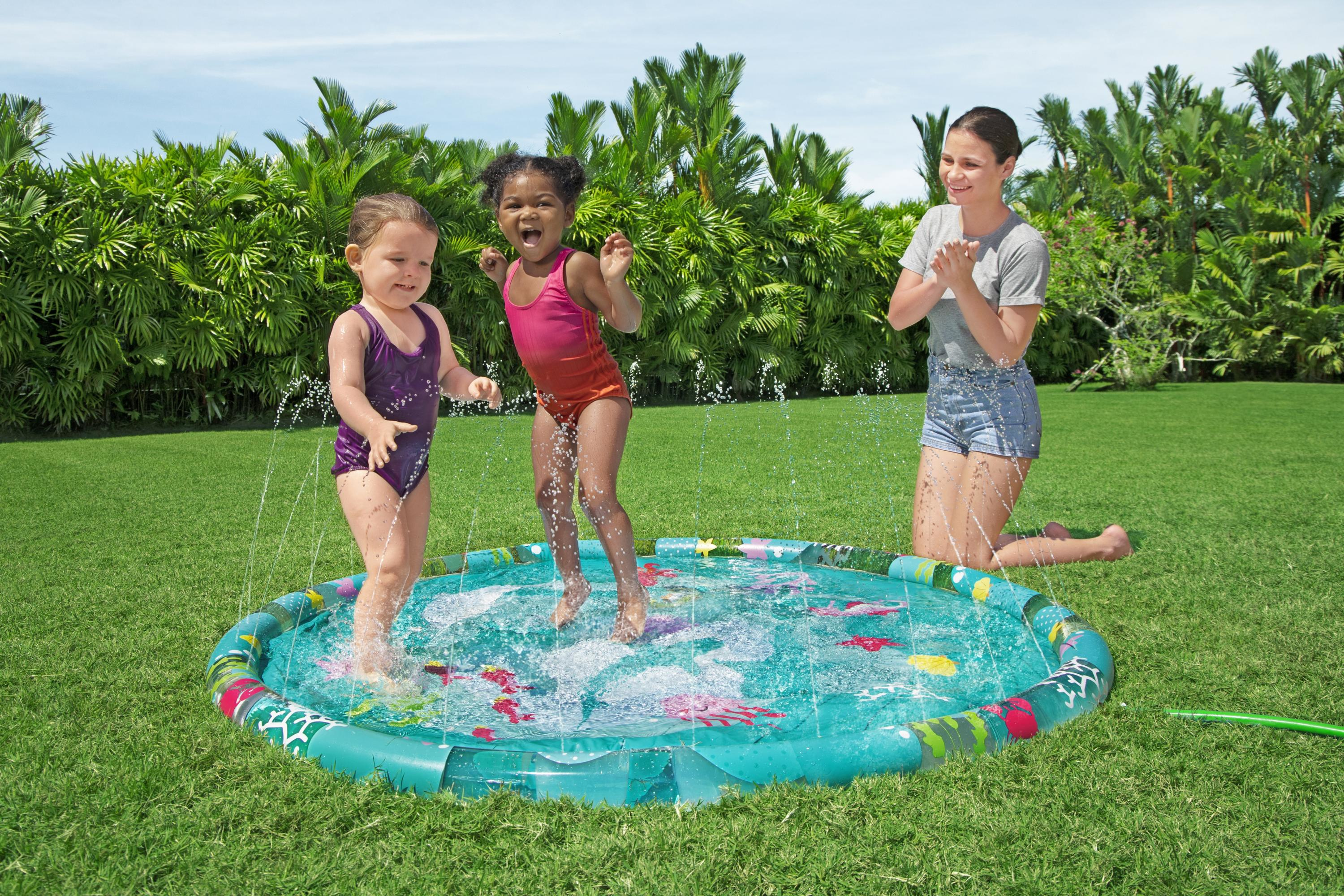 Dos niñas pequeñas juegan en una piscina inflable azul con chorros de agua, mientras una mujer adulta se arrodilla cerca, sonriendo.