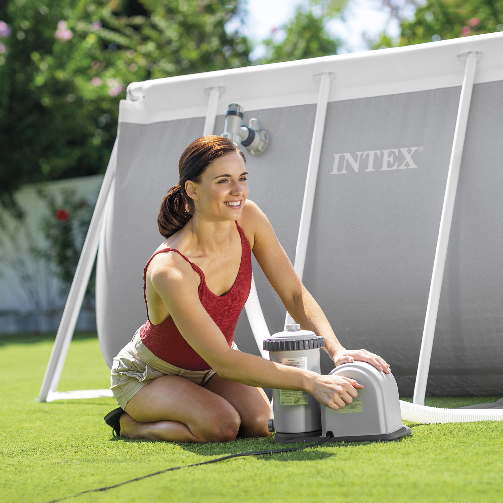 Donna in costume rosso e pantaloncini si inginocchia vicino al filtro piscina Intex grigio.