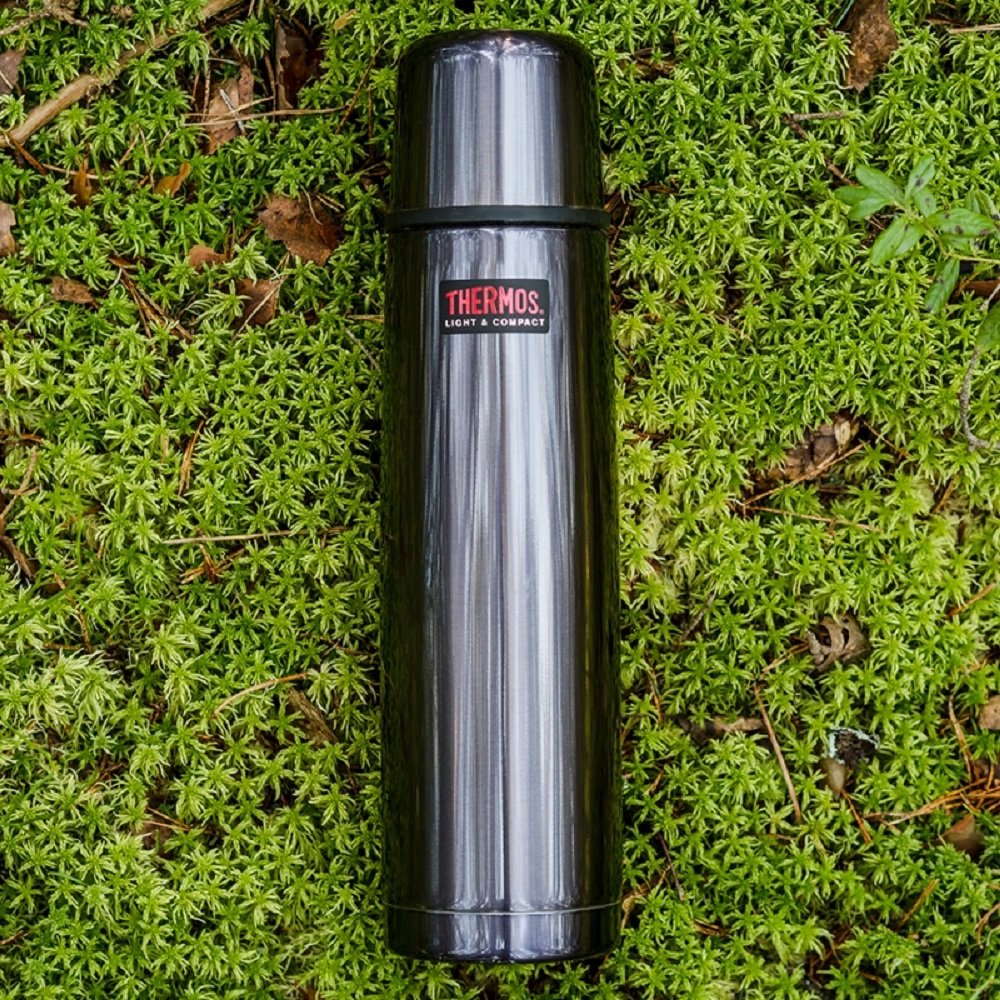 Een zilveren thermoskan met rood Thermos-logo op groen mos.