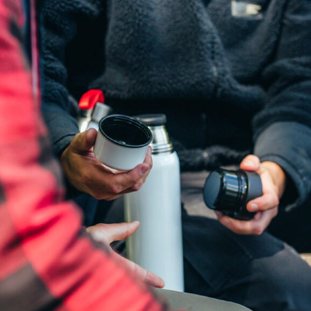 Twee personen houden een witte thermoskan en twee deksels vast. Een deksel is wit, de ander zwart.