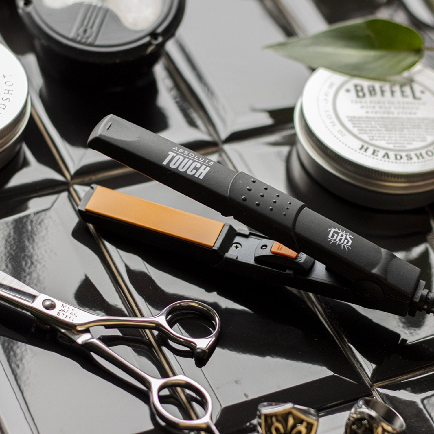 A flat iron, scissors, and two cans on a shiny black surface.