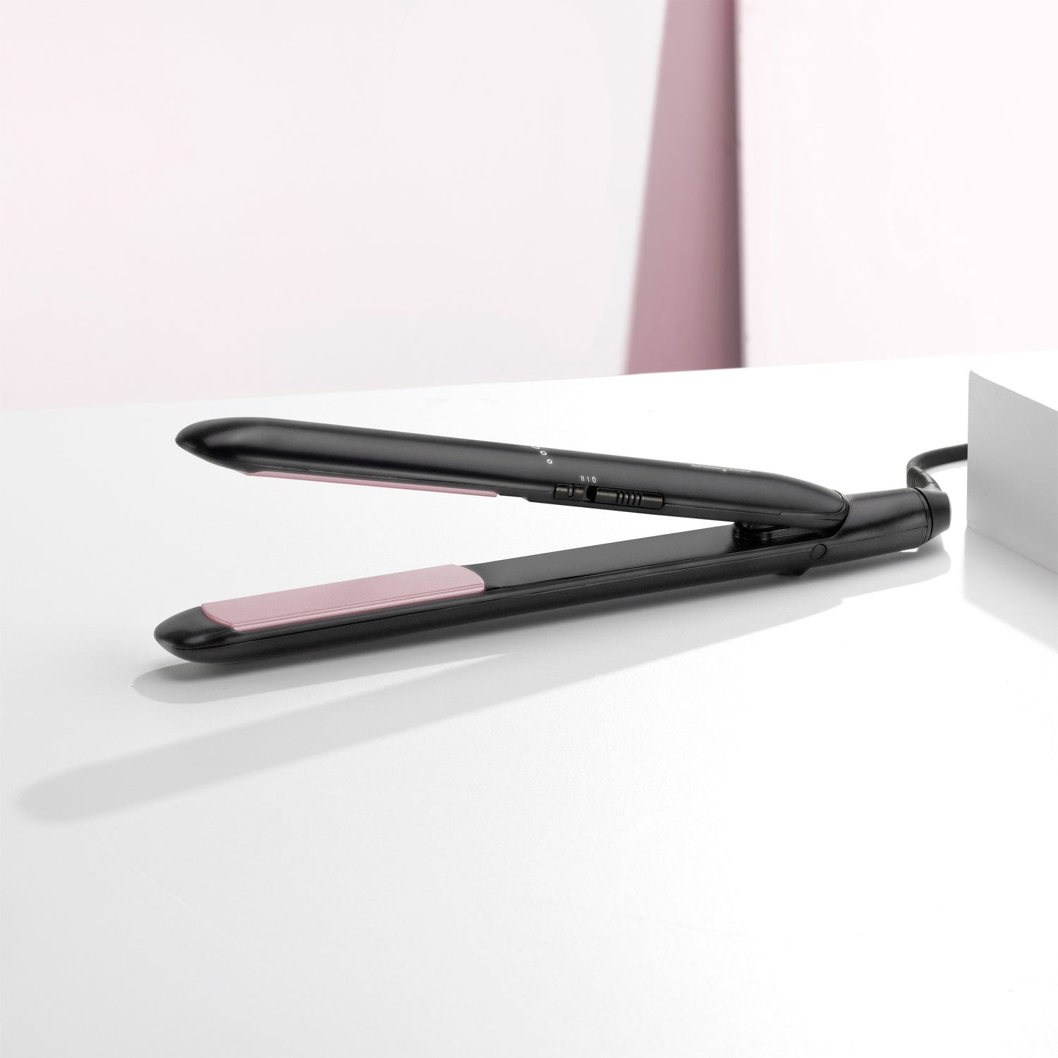 A black hair straightener with pink plates lies open on a white surface.