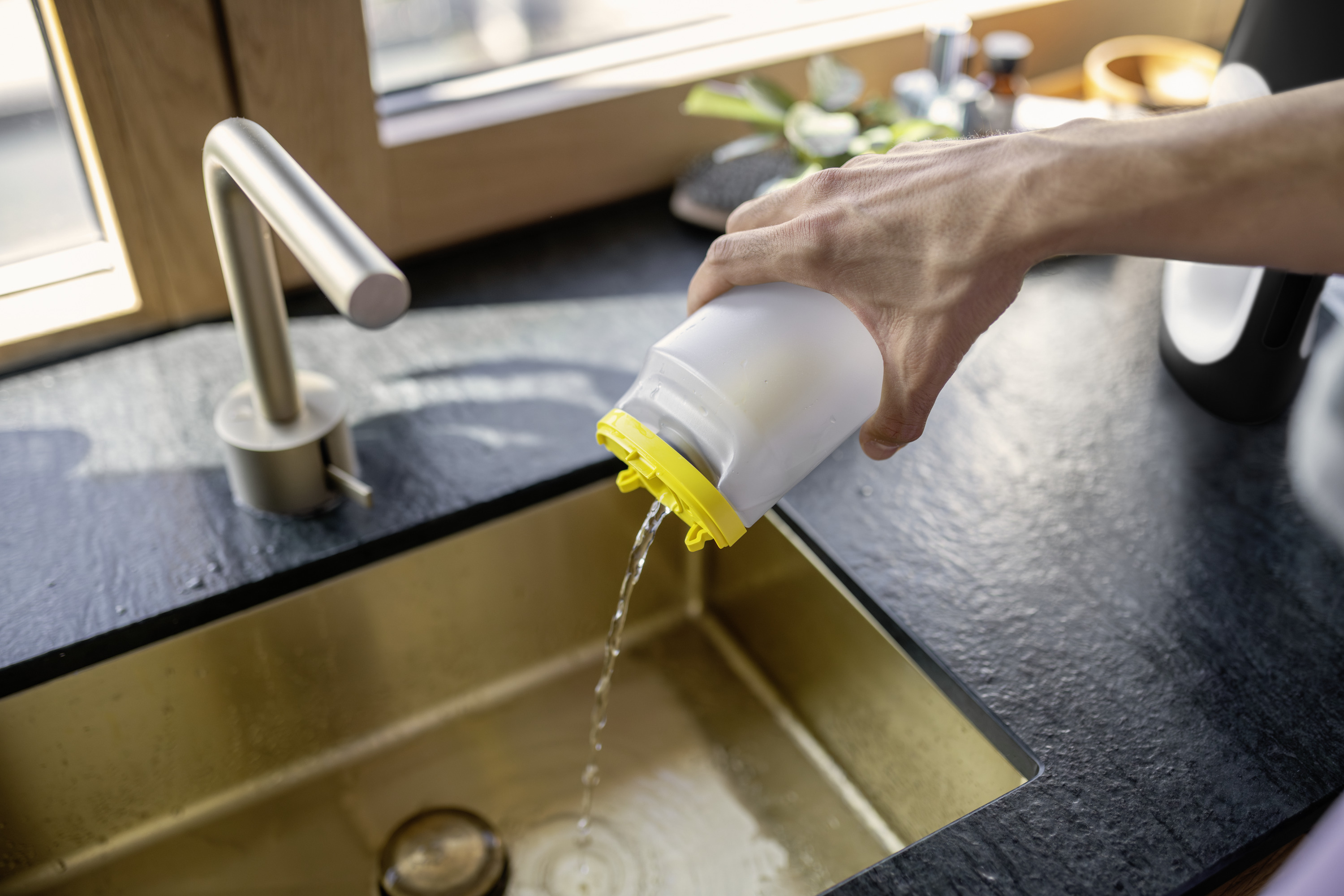 Wasser wird aus einem weißen Behälter mit gelbem Deckel in ein goldenes Spülbecken gegossen.