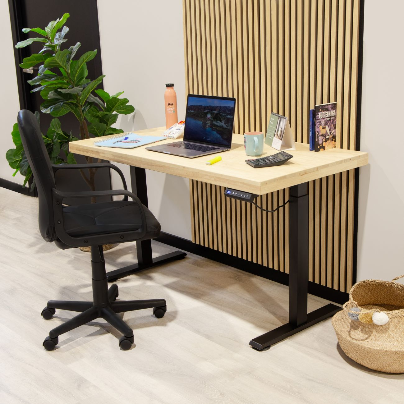 Un escritorio regulable en altura con portátil, silla, planta y cesta.