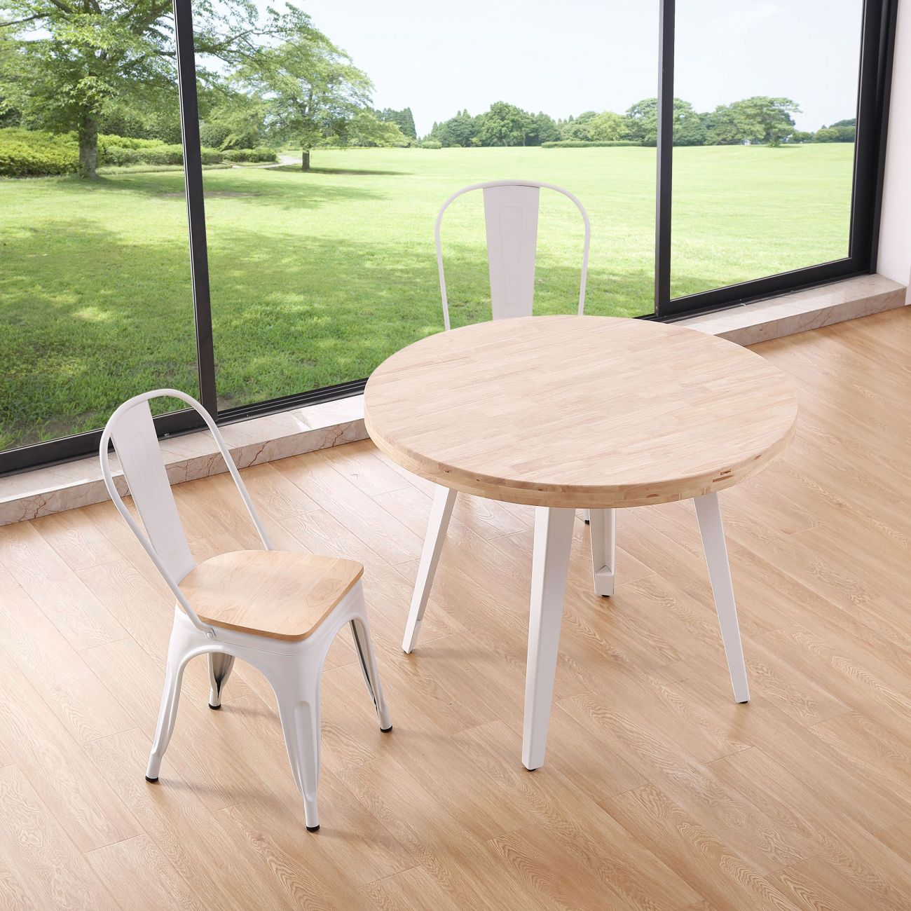 Mesa redonda de madera con sillas blancas frente a una ventana con vistas a un césped verde.