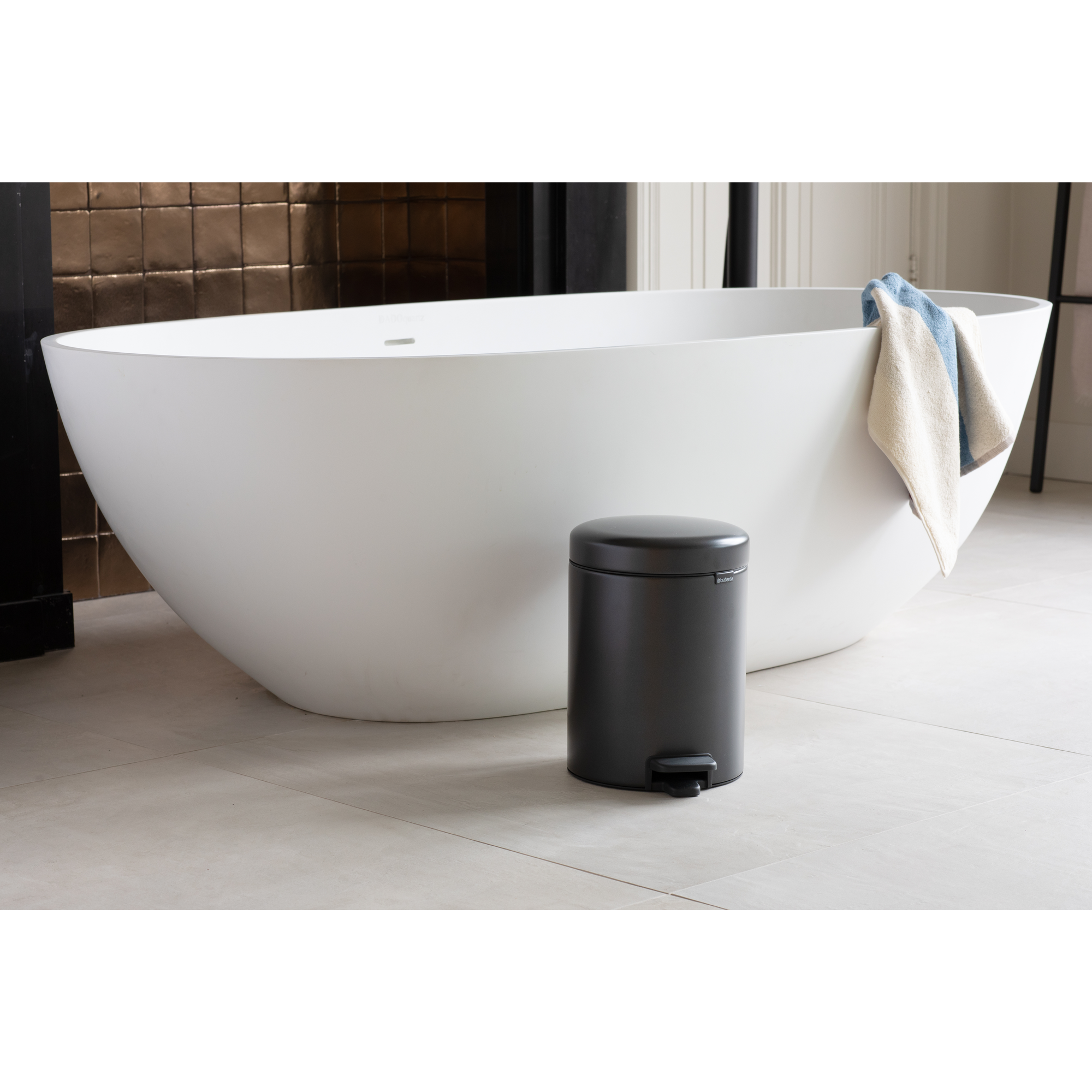 Un cubo de basura gris oscuro frente a una bañera blanca con toallas.