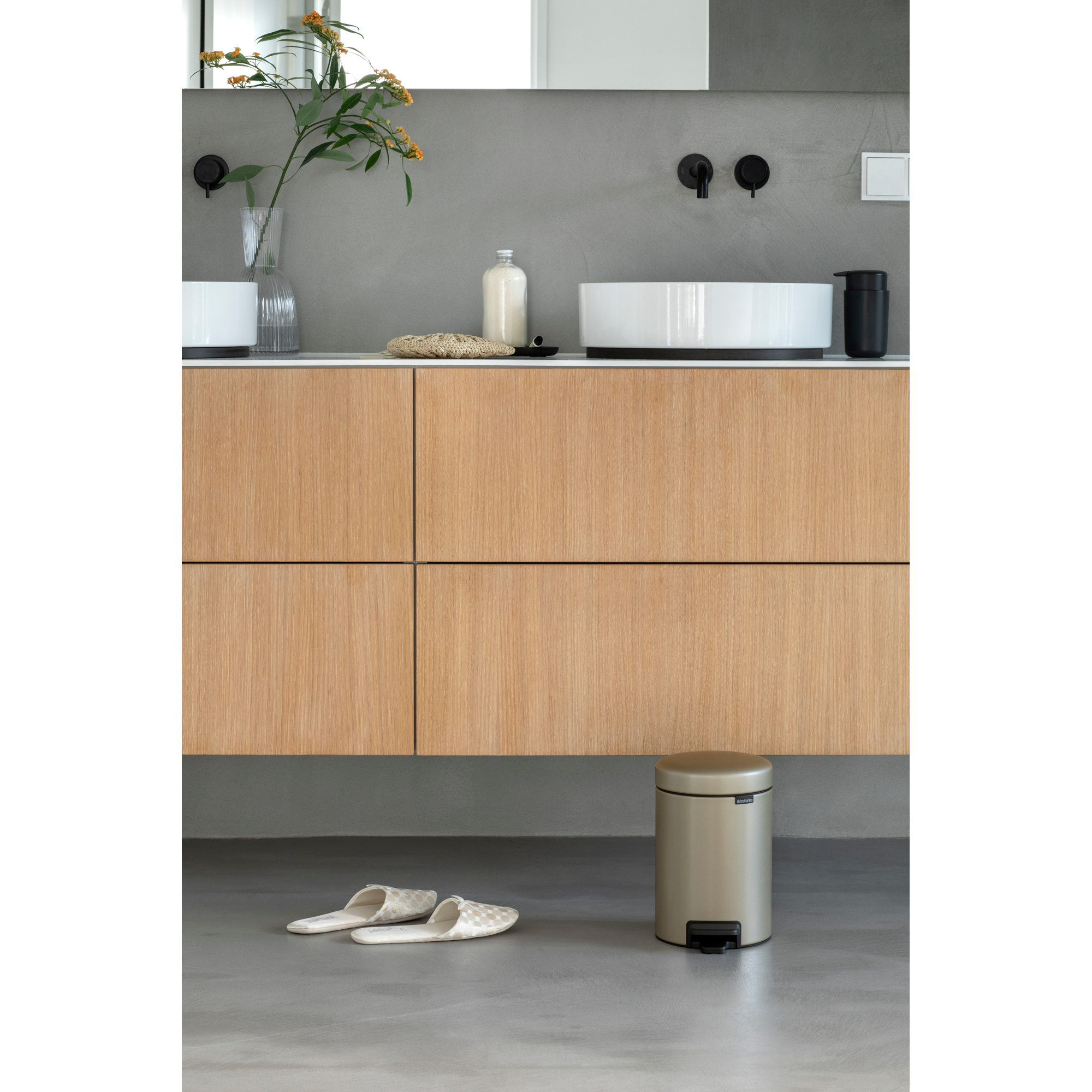 Baño con mueble de madera, lavabo, cubo y zapatillas.